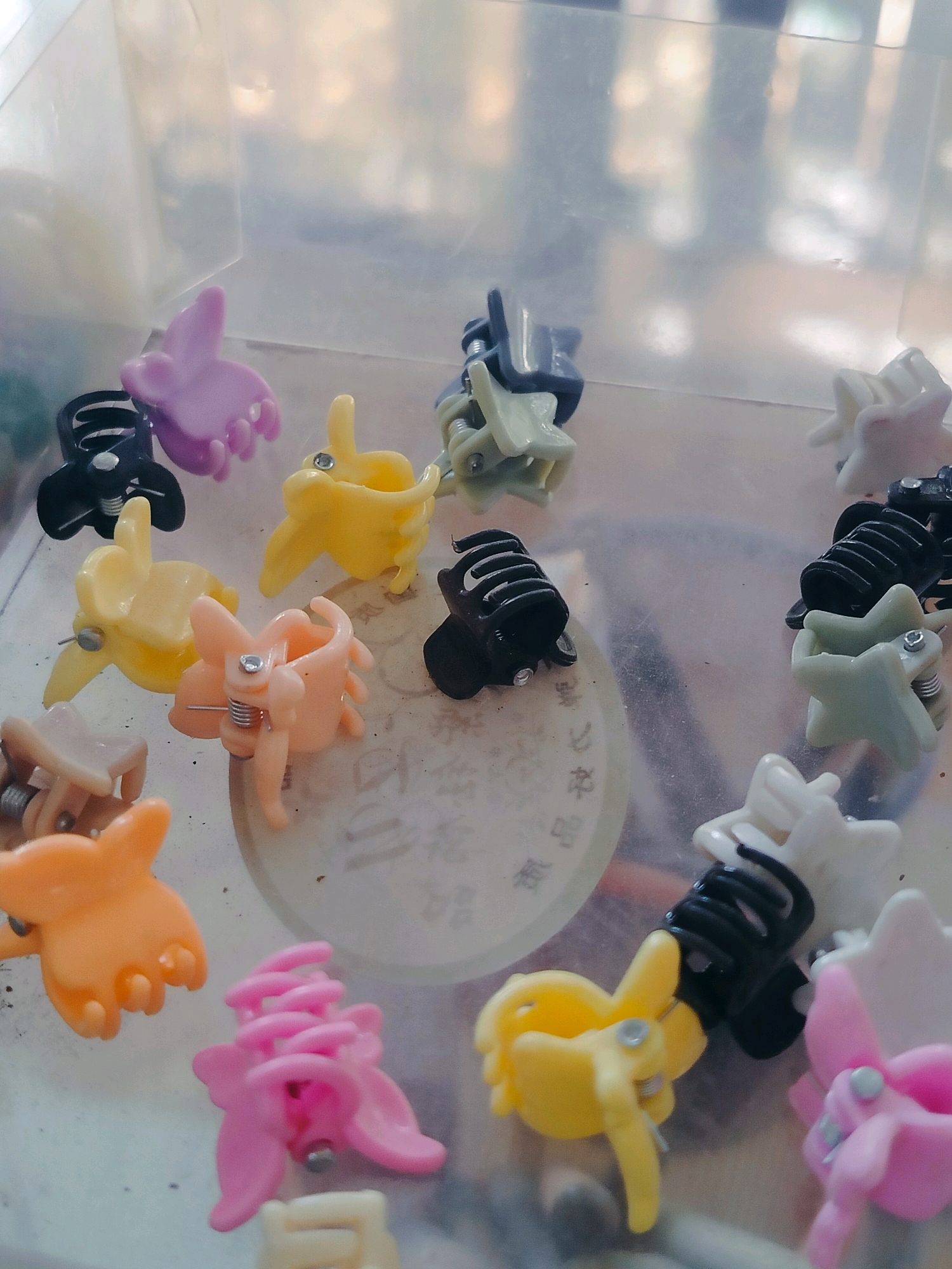 A group of colorful plastic hair clips are scattered inside a clear plastic container. The clips are in various shapes and colors, including purple, yellow, pink, orange, black, and light green. Some of the clips are shaped like flowers, while others are more abstract designs. The container is clear and slightly scratched, with a faint circular pattern visible on the bottom of the container. The background is blurred and out of focus, suggesting an indoor setting. The image appears to be a still life photograph. There are no people or animals present. The lighting is bright and diffused, casting soft shadows on the clips. The location is not discernible from the image content.