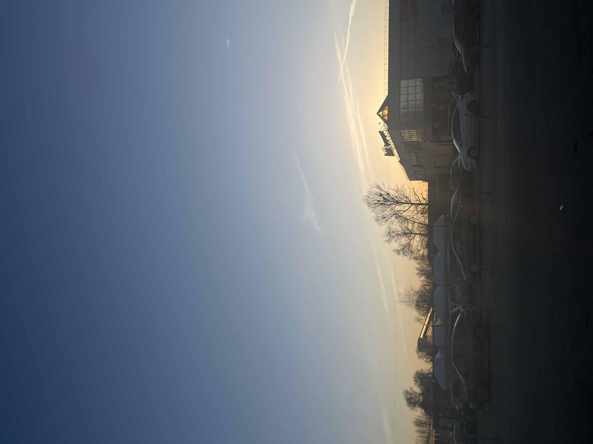 This image depicts an outdoor commercial scene in Klin, Russia, at either sunrise or sunset, evidenced by the low sun angle and golden light on the horizon. The sky transitions from a deep blue overhead to a lighter blue and then a golden-orange near the horizon, marked by two intersecting contrails. In the foreground, a dark, indistinct area, possibly a road or pavement, features a low horizontal barrier. The midground contains a line of various vehicles, mostly sedans and possibly an SUV, rendered as dark silhouettes against the bright horizon, suggesting a street or parking lot. No specific actions or interactions between people or vehicles are discernible.
On the right side of the background stands a multi-story commercial building with light-colored facades, numerous windows, and a prominent dark-framed pyramidal glass roof structure. Signage on this building clearly identifies it as "СИТИЛИНК" (CITILINK) and "DNS," indicating electronics retail stores. To the left of this building, behind the vehicles, several white tent-like structures, some with visible snow accumulation, are present, suggesting temporary outdoor stalls or shelters. Further left and behind these structures, additional indistinct buildings and bare deciduous trees are visible, characteristic of late autumn or winter. The overall environment is an urban or suburban commercial district within Klin, Russia.