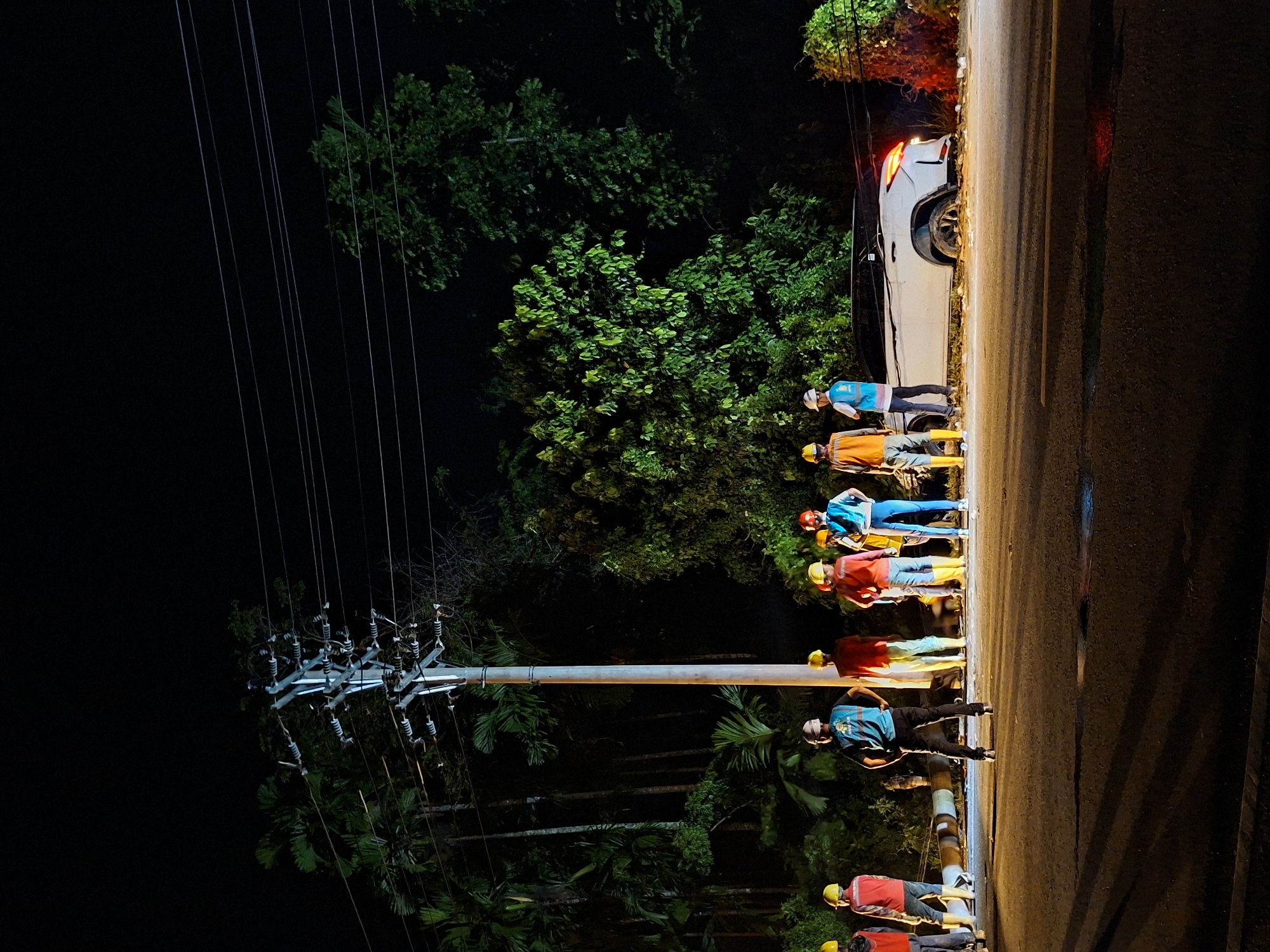 At night in Teungoh Baro, Indonesia, a group of seven workers stand on the side of a road next to a white car with its rear lights illuminated. The workers are wearing various combinations of safety vests, helmets, and long-sleeved shirts. A utility pole with power lines runs parallel to the road, near lush green trees and foliage. The scene is illuminated by artificial lighting, casting shadows on the road and the surrounding environment.