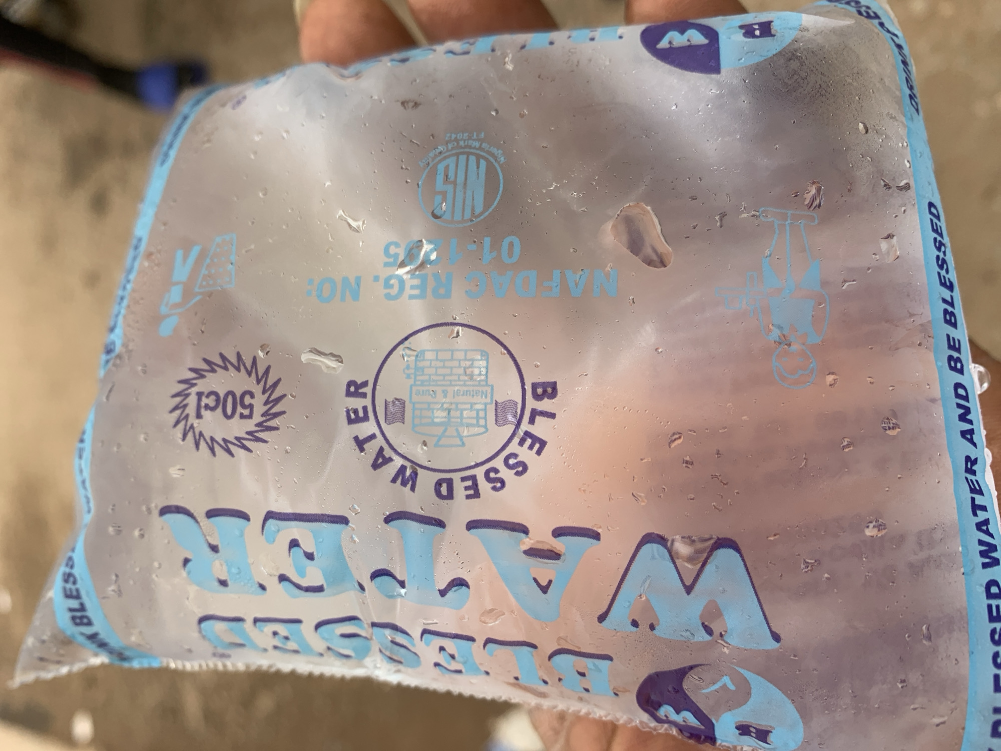 The image shows a clear plastic sachet of "BLESSED WATER" being held by a hand. The sachet is filled with water and has blue lettering and graphics printed on it. Prominently displayed is the word "WATER" in large, light blue capital letters. Above it, "BLESSED" is also printed in large, light blue capital letters, vertically.
The sachet also features a circular logo with the text "BLESSED WATER" around it and an image of a building inside. To the right of this logo, text reads "NAFDAC REG. NO: 01-1205" and a circular "NIS" logo. The volume "50cl" is also visible in a starburst graphic.
The sachet is slightly condensation-covered, indicating it is likely cold. The background is blurred, but suggests an outdoor setting with some muted colors. The hand holding the sachet appears to be a lighter skin tone. The presence of the NAFDAC registration number suggests the product is from Nigeria. While the image itself does not explicitly show the city of Shagamu, Nigeria, such sachets of water are commonly sold in various cities across Nigeria, including Shagamu.