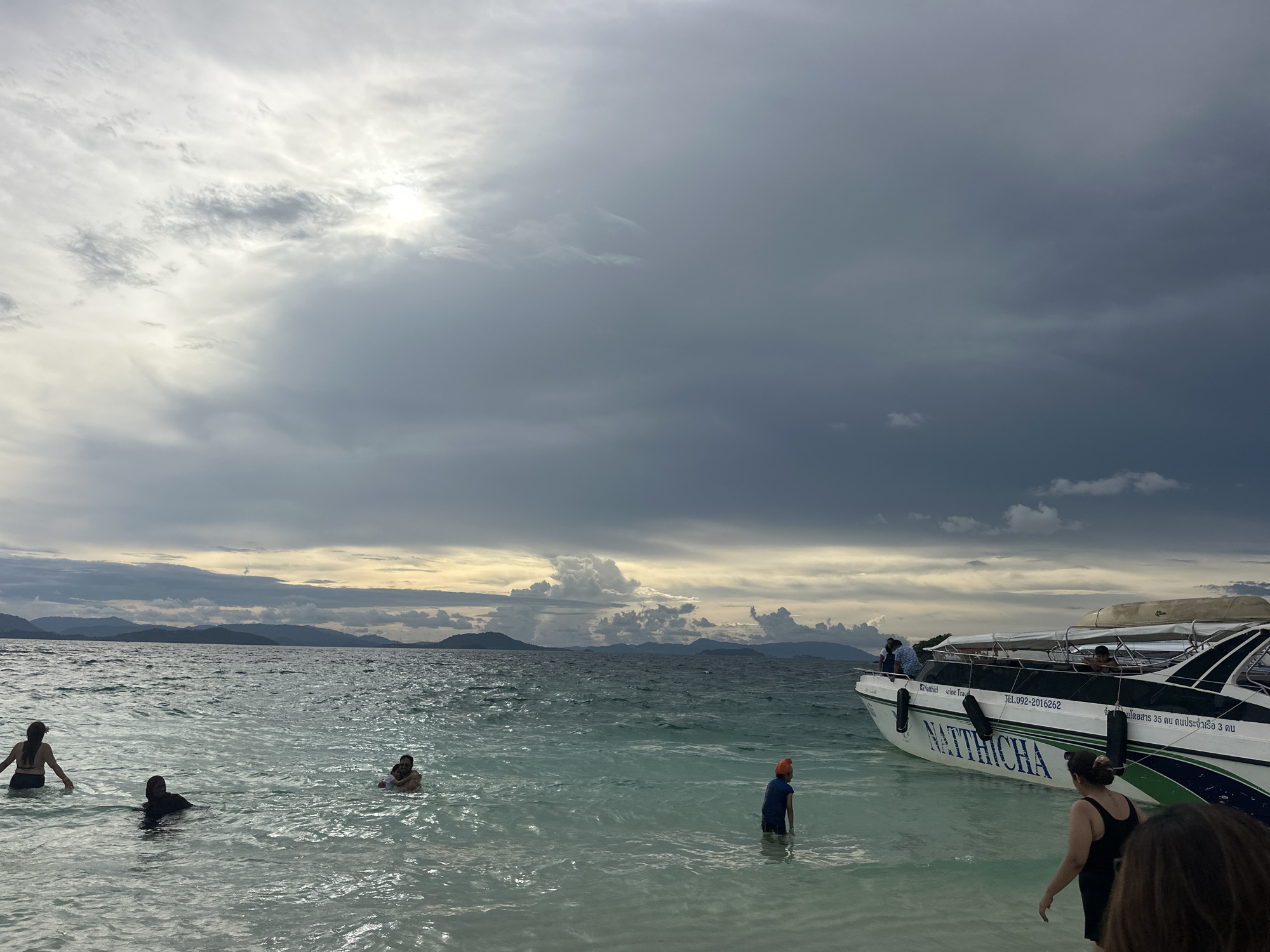 The image depicts a cloudy day at a beach in Tambon Phru Nai, Thailand. In the water, several individuals are visible. To the left, a person with long dark hair is wading in the ocean. Further into the water, another person in a black top is partially submerged. To the right of these individuals, a person wearing an orange hat and a blue shirt stands in the water.
On the right side of the frame, a white speed boat named "NATTHICHA" is docked. The boat has several passengers and crew members visible. A woman in a black tank top is walking towards the boat from the water. In the foreground, a portion of another person's head and shoulder is visible.
The ocean water is a clear turquoise color, with visible ripples and waves. In the background, a chain of islands or mountains with lush green vegetation can be seen against the horizon. The sky is dominated by heavy, dark clouds, with patches of sunlight breaking through in the upper left portion of the image, creating a dramatic and moody atmosphere. The overall scene suggests a tropical beach setting with overcast weather.