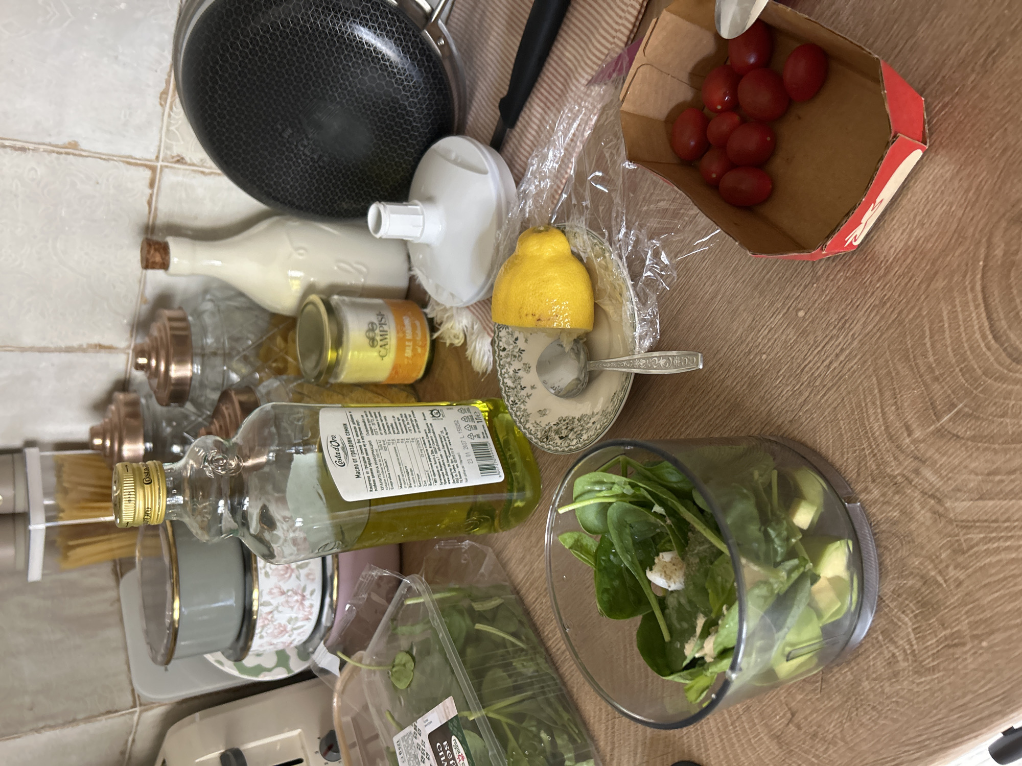 The image depicts a kitchen counter with various ingredients and cooking utensils. In the foreground, a clear plastic container holds spinach leaves and avocado pieces, presumably being prepared for a meal. Next to it, a halved lemon sits on a small plate with a spoon, while a separate box contains a cluster of cherry tomatoes. A bottle of olive oil, a jar of what appears to be seasoning or spices, and a white ceramic jug are also visible. In the background, a pan and several storage containers, including one filled with pasta, are arranged. The overall setting suggests a home kitchen environment. There is no information in the image to suggest a specific location in Plovdiv, Bulgaria.