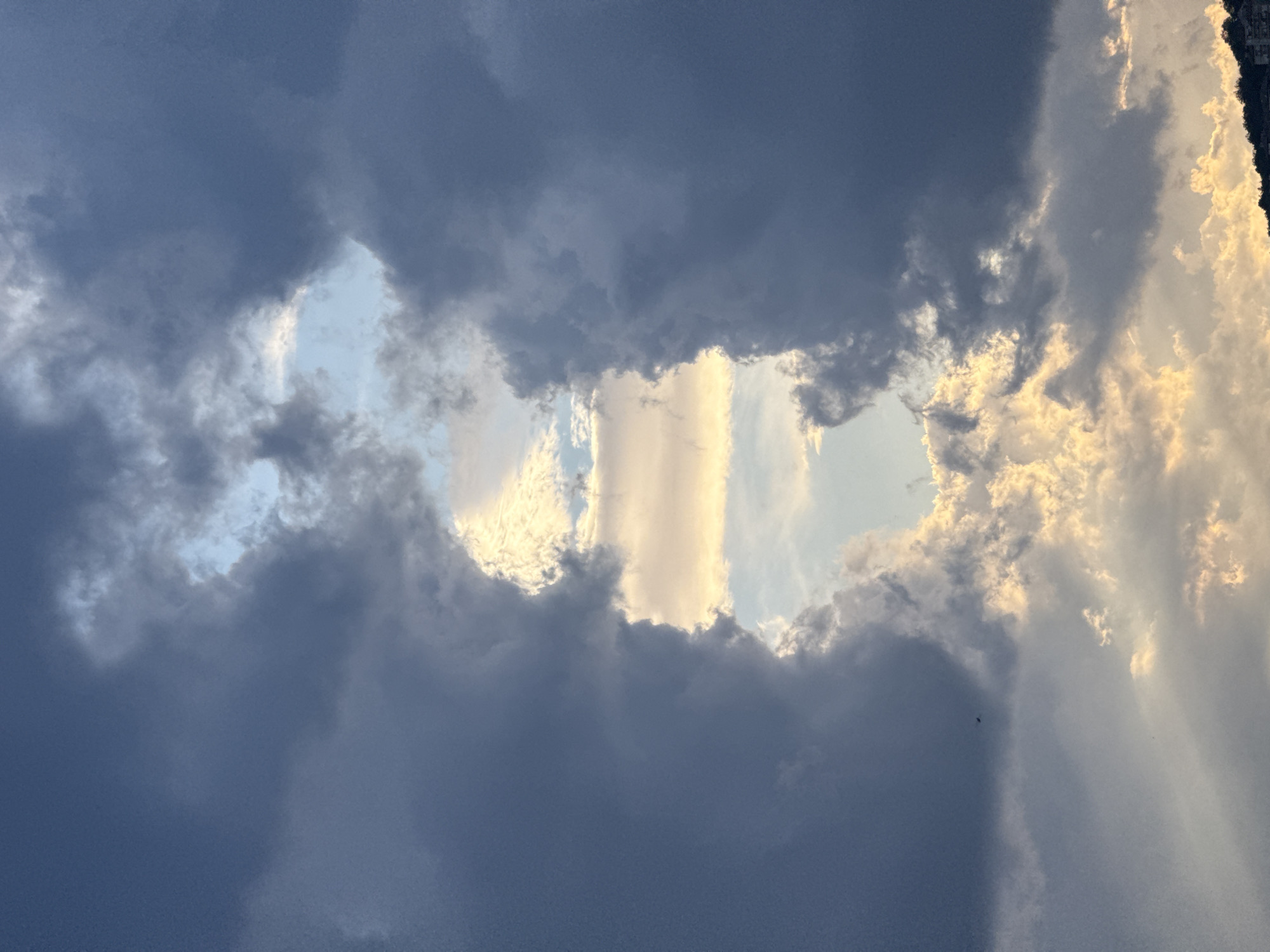 The image shows a view of the sky with dark, heavy clouds covering most of the frame. There are openings in the clouds, revealing patches of light blue sky and illuminated clouds with a golden hue. The image has been rotated 90 degrees counterclockwise. There are no visible people, subjects, objects, actions, or interactions within the scene. The location in the city of Veliko Tarnovo, Bulgaria is not discernable from the image.