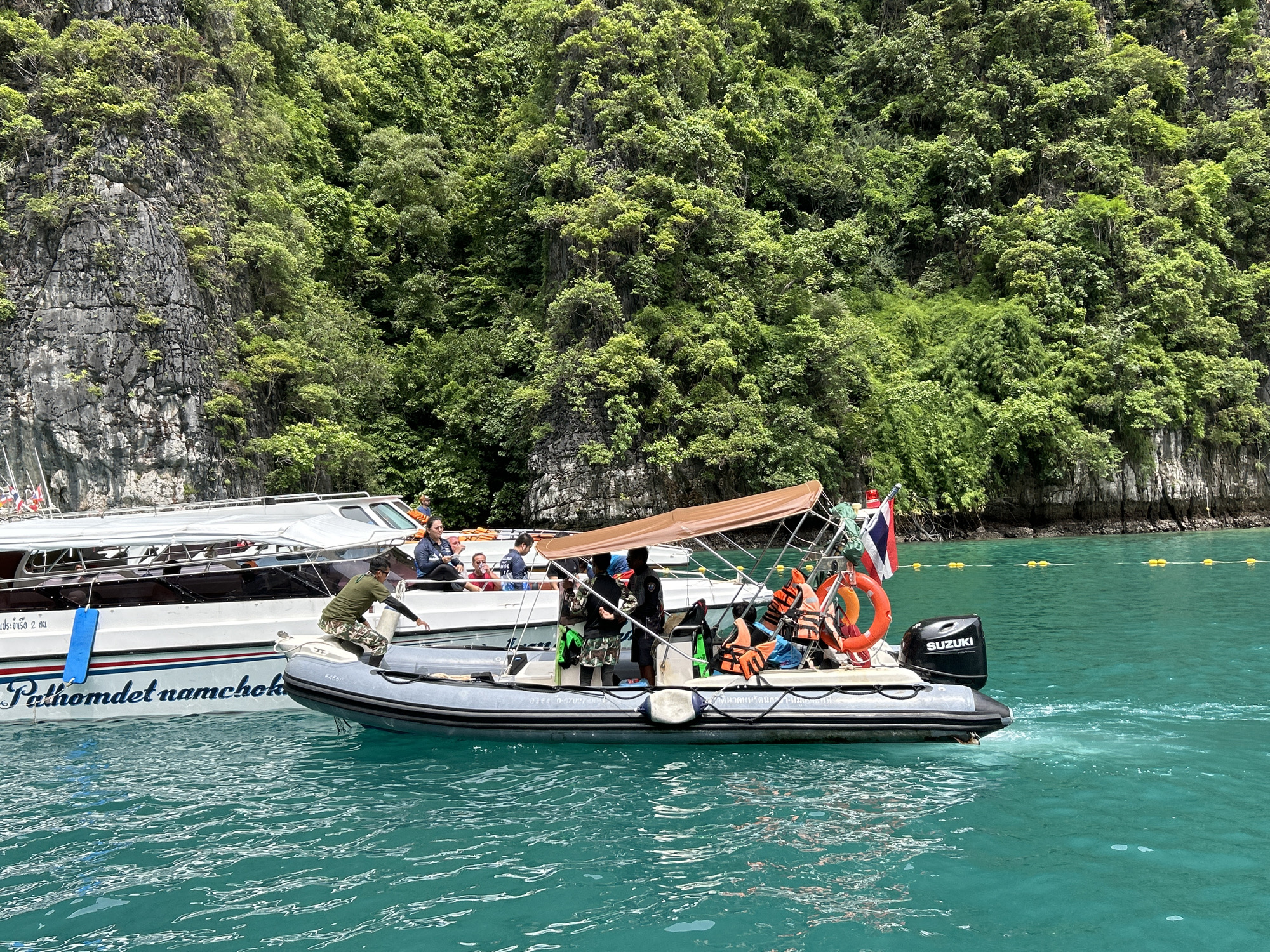 A grey inflatable dinghy with an outboard motor is positioned in turquoise water next to a larger white passenger boat. The dinghy has a brown canopy overhead and several people wearing life vests are seated inside. A man in a green shirt and camouflage shorts is standing in the dinghy. The larger boat has "Pathomdet namchok" written on its side in blue lettering. In the background, a lush green, forested cliff face rises from the water. The scene appears to be in Ao Nang, Thailand, a popular tourist destination known for its limestone karst formations and clear waters.