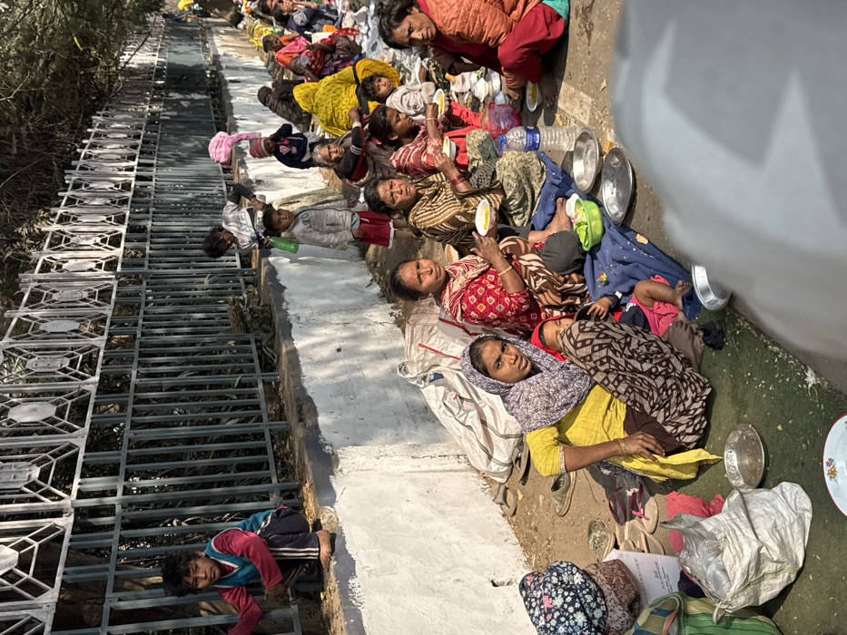 A group of women and children are lying on the ground beside a painted white wall. They are holding plates of food and some are eating. A metal grate runs along the other side of the wall. A child is reaching through the grate. The people are wearing various clothing and are lying on blankets and bags. There are water bottles and sandals scattered around.