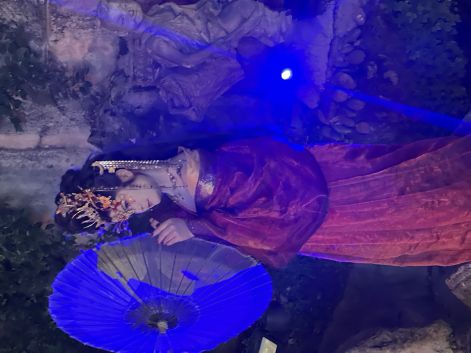 The image displays an individual in elaborate traditional Chinese attire within a dimly lit, staged environment, specified as Yuzhong District, China. The central figure, appearing to be an adult female, is positioned in a three-quarter profile facing left. She holds a large, open, deep blue traditional Chinese umbrella with both hands, her right hand gently touching its edge. Her costume consists of a flowing, sheer magenta-red outer robe over an iridescent gold undergarment and a red skirt. Her dark hair is styled upwards, adorned with an ornate golden headpiece featuring intricate designs and dangling chains. Facial makeup is prominent, including white foundation, red markings on the forehead and cheeks, and defined eyes. In the right background, partially obscured by blue illumination, are several carved stone or cement statues, one depicting a smiling seated figure. The scene is predominantly illuminated by a strong blue light source on the right, casting a vibrant blue hue over the background and the umbrella, which contrasts with the rich red and gold tones of the costume. Green foliage and stone textures are visible in both the background and foreground.