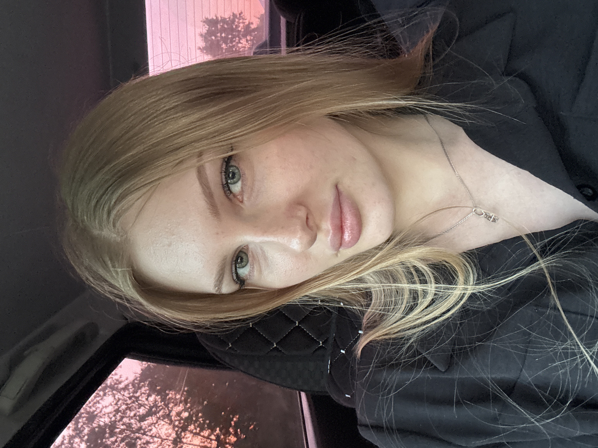A close-up, slightly angled photograph of a young woman with light blonde hair inside a vehicle. The woman has fair skin and visible freckles, with her face turned toward the camera. She wears a dark-colored, collared shirt and a silver necklace. Outside the car window, a blurred background suggests a tree and a pinkish-purple sky. The interior of the vehicle includes a black quilted material, likely a car seat. The image suggests a moment captured in transit, with soft lighting and a relaxed pose. It is not possible to confirm a location in the city of Kondratovo, Russia without further information.