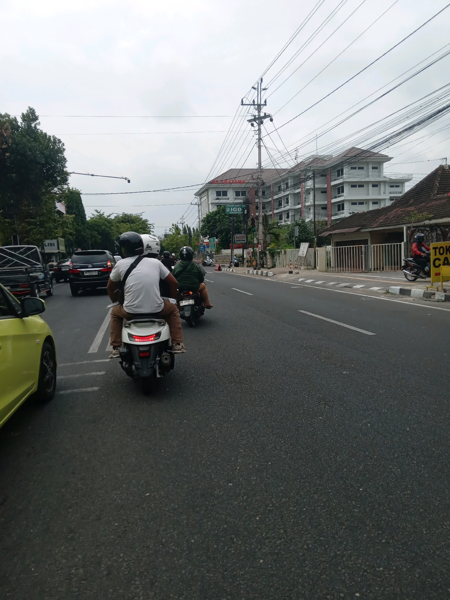 A street scene in Yogyakarta, Indonesia, shows a moderate flow of traffic under an overcast sky. The main focus is on two individuals riding a scooter from a rear-facing perspective. The rider wears a white t-shirt and brown pants, with a black helmet. The passenger, behind the rider, wears a green jacket and a black helmet. They are riding on the right side of a paved road.
To the left of the scooter, a portion of a yellow car is visible. Further in the background, a black SUV and other vehicles are present. The road is marked with white lines. On the right side of the road, a building with multiple floors, appearing to be a hospital or clinic, is visible. A sign with Indonesian text is present near the building entrance. Adjacent to the hospital is a smaller building with a tiled roof and a white fence. Several utility poles with numerous wires are prominent in the background, extending across the frame. Trees line the left side of the road. The overall impression is a typical urban street in Indonesia during daytime.