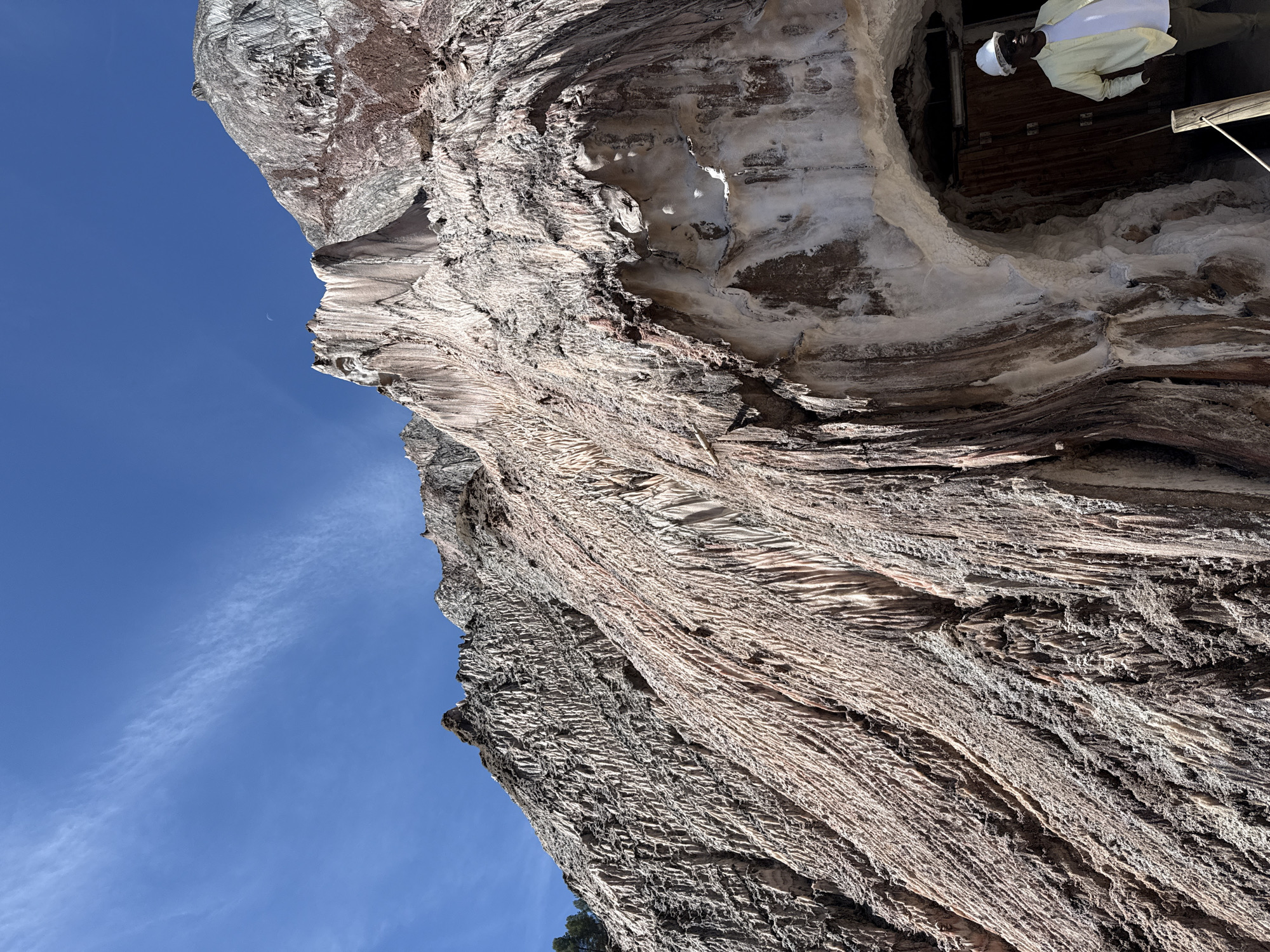 In Cardona, Spain, a man wearing a white hard hat and a light yellow long-sleeved shirt stands to the right of the frame, looking into an arched opening in a massive, multi-colored salt mountain. The mountain's surface is textured with vertical striations and exhibits a gradient of colors ranging from white and pink to reddish-brown. The opening appears to be an entrance to a mine or underground area, with wooden doors visible inside. The sky above is a clear, bright blue with thin, wispy clouds.