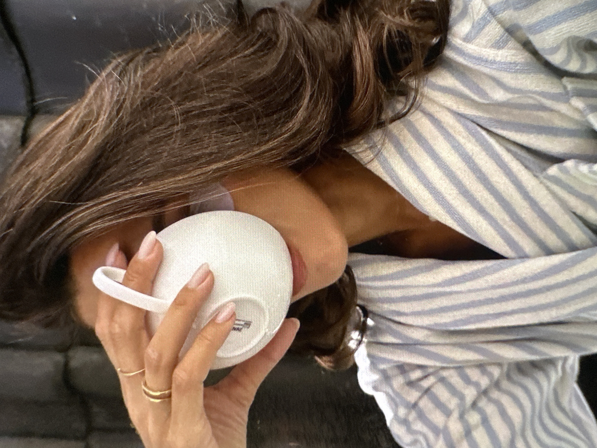 A woman with long brown hair is lying down, partially covered by a light blue and white striped garment. She's holding a white mug to her lips with her right hand. The mug appears to be empty. She has two gold rings on her fingers. The background is dark and out of focus. The image's location and city are not discernible from the visual information.