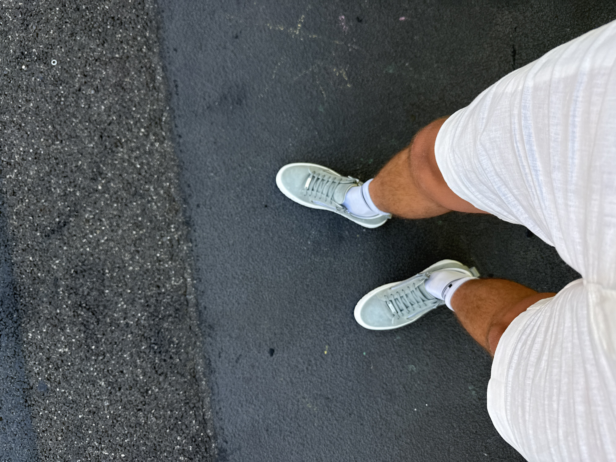 A first-person perspective shot shows a person's legs and feet standing on a wet asphalt surface. The person is wearing white shorts and light grey sneakers with white socks. The asphalt has a rough texture and appears to be recently wet, with some lighter patches and subtle reflections. The scene is outdoors, with no other people or distinct landmarks visible. The location is Scarperia, Italy.