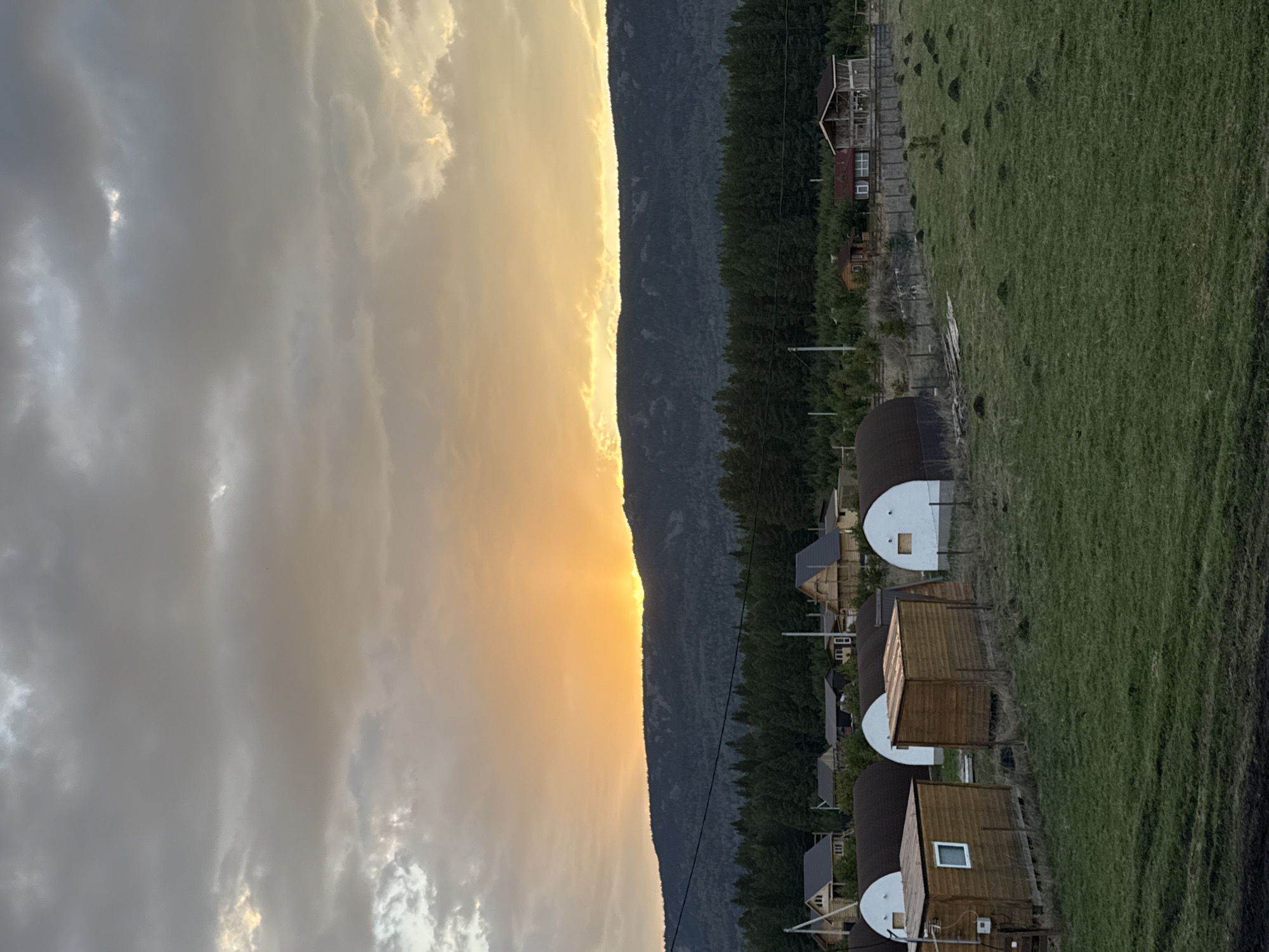 The image shows a landscape in Tyulyuk, Russia, featuring a line of small wooden cabins with domed structures along a green field, bordering a dense forest. A mountain range rises in the background, illuminated by a bright, possibly setting sun. The sky is filled with gray and white clouds. There are no people visible.