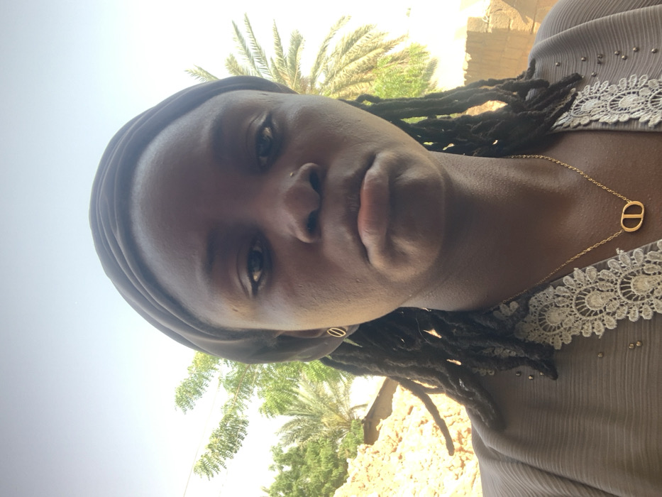 A close-up portrait shot depicts an individual with dark skin, wearing a dark grey head covering that reveals dreadlocks beneath. The individual has a small gold-colored stud piercing in the right nostril and wears small gold-colored circular stud earrings. A light grey or brown garment with a white lace and beaded neckline is visible, adorned with a gold-colored chain necklace featuring an oval "CD" pendant. The individual looks directly at the camera with a neutral expression, lips slightly pursed. The background shows a bright, clear sky above various green foliage, including palm fronds and leafy trees. A light-colored, possibly mud-brick or sand-colored structure is partially visible on the left side of the background. The scene is located in Bojude, Nigeria.