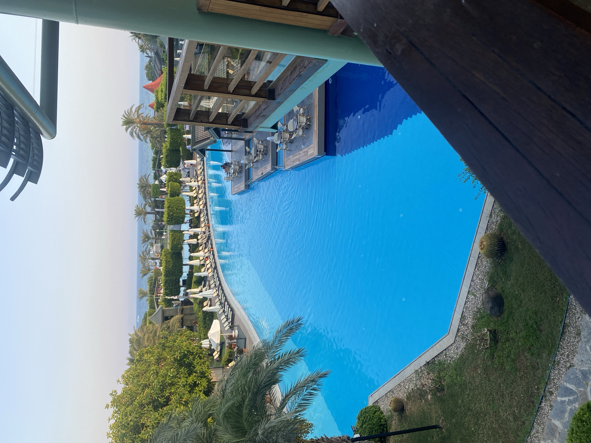 The image shows a large, bright blue swimming pool at what appears to be a resort in Aksu, Turkey. The pool is a prominent feature, with loungers and umbrellas set up along its edge. Several floating platforms with tables and chairs are situated in the middle of the pool, suggesting an outdoor dining or seating area. Palm trees and other lush greenery surround the pool, and the sea is visible in the background, with a clear sky above. Parts of a wooden structure, likely a balcony or bridge, are in the foreground and right side of the image. The scene depicts a tranquil resort environment, likely during daylight hours.