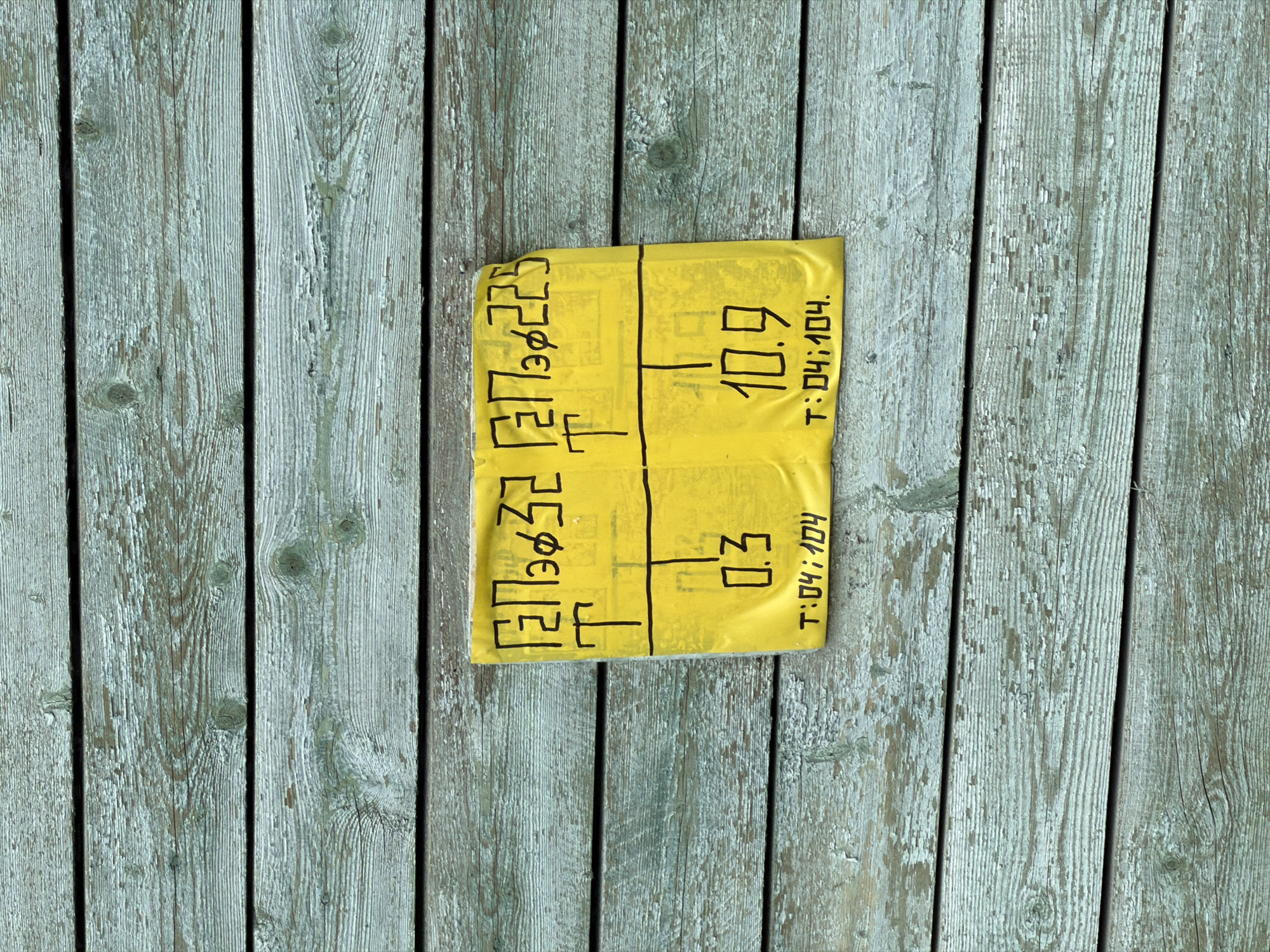 A rectangular piece of yellow paper with hand-drawn black markings lies on a weathered wooden surface composed of light greenish-gray planks. The paper is divided into two columns and two rows. The left side has illegible symbols. The right top corner says "10.9" and the right bottom corner says "0.3". The paper also contains "T: 04:104."
The image was taken in Strelna, Russia.