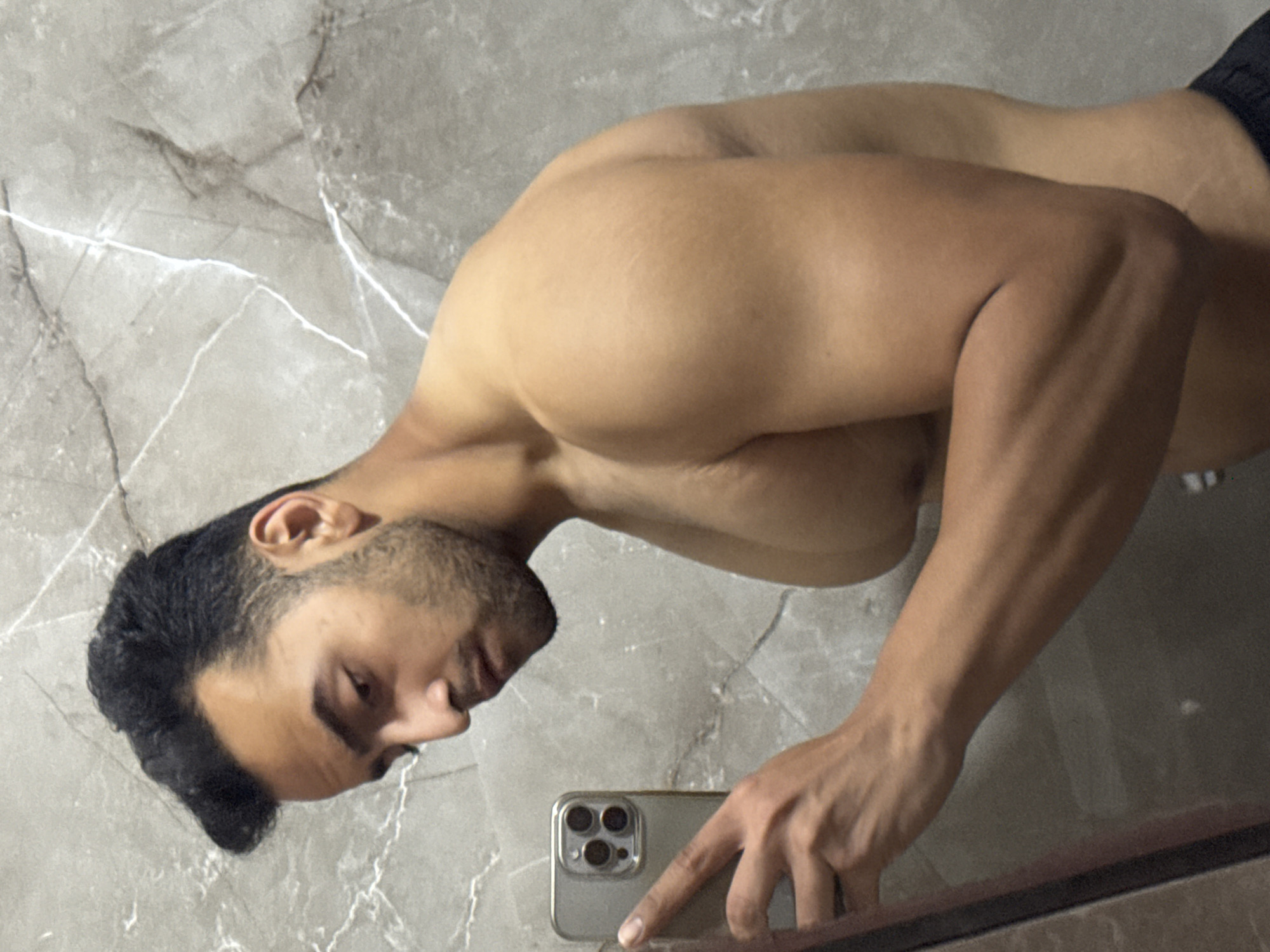 A shirtless man is taking a selfie in front of a bathroom mirror with gray marble walls. The man is holding a smartphone with his right hand. He has dark hair and a trimmed beard. The location is likely in Kolkata, India.