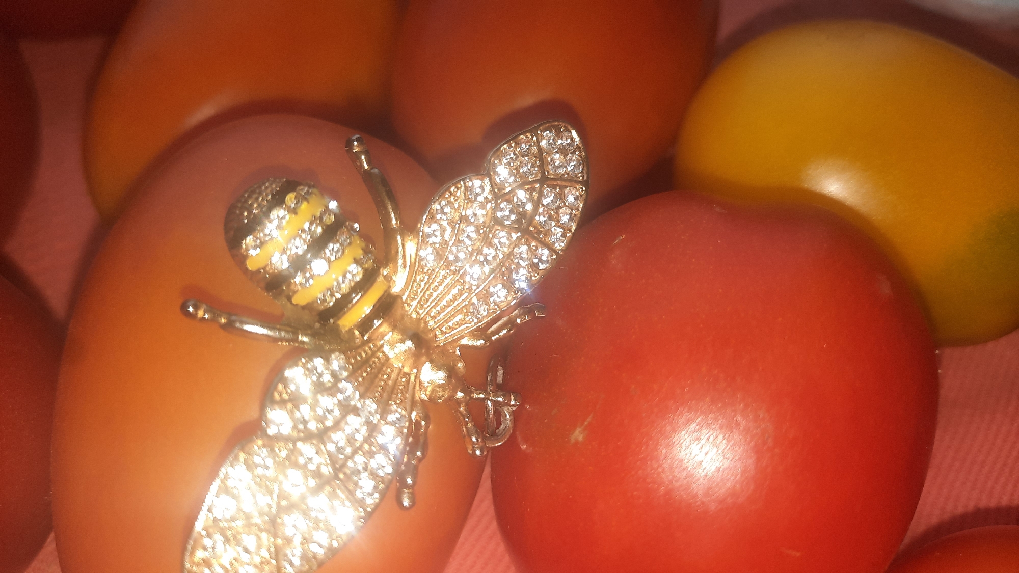 A close-up, overhead shot shows a decorative bee brooch placed on a group of tomatoes. The brooch is gold with clear rhinestones and yellow stripes on its abdomen. It rests on an orange tomato, with a red tomato visible to its right and other tomatoes, some yellow and some orange, in the background. The tomatoes appear to be ripe. The scene is indoors, with a soft pink surface beneath the tomatoes. There is no indication of the location being in the city of Шклоў, Belarus.