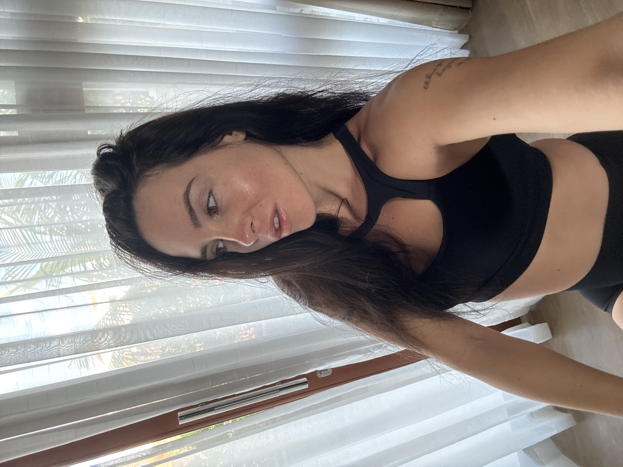 A woman with long, dark hair and fair skin is shown from the chest up. She is wearing a black sports bra and black shorts. She has a tattoo on her left arm. She is looking to the right of the frame with her mouth slightly open. The background consists of sheer white curtains that allow sunlight to filter through, revealing the silhouettes of palm trees. A portion of a wooden frame and a vertical metal pole are visible on the left side of the image. The scene appears to be indoors, possibly in a room with a large window.