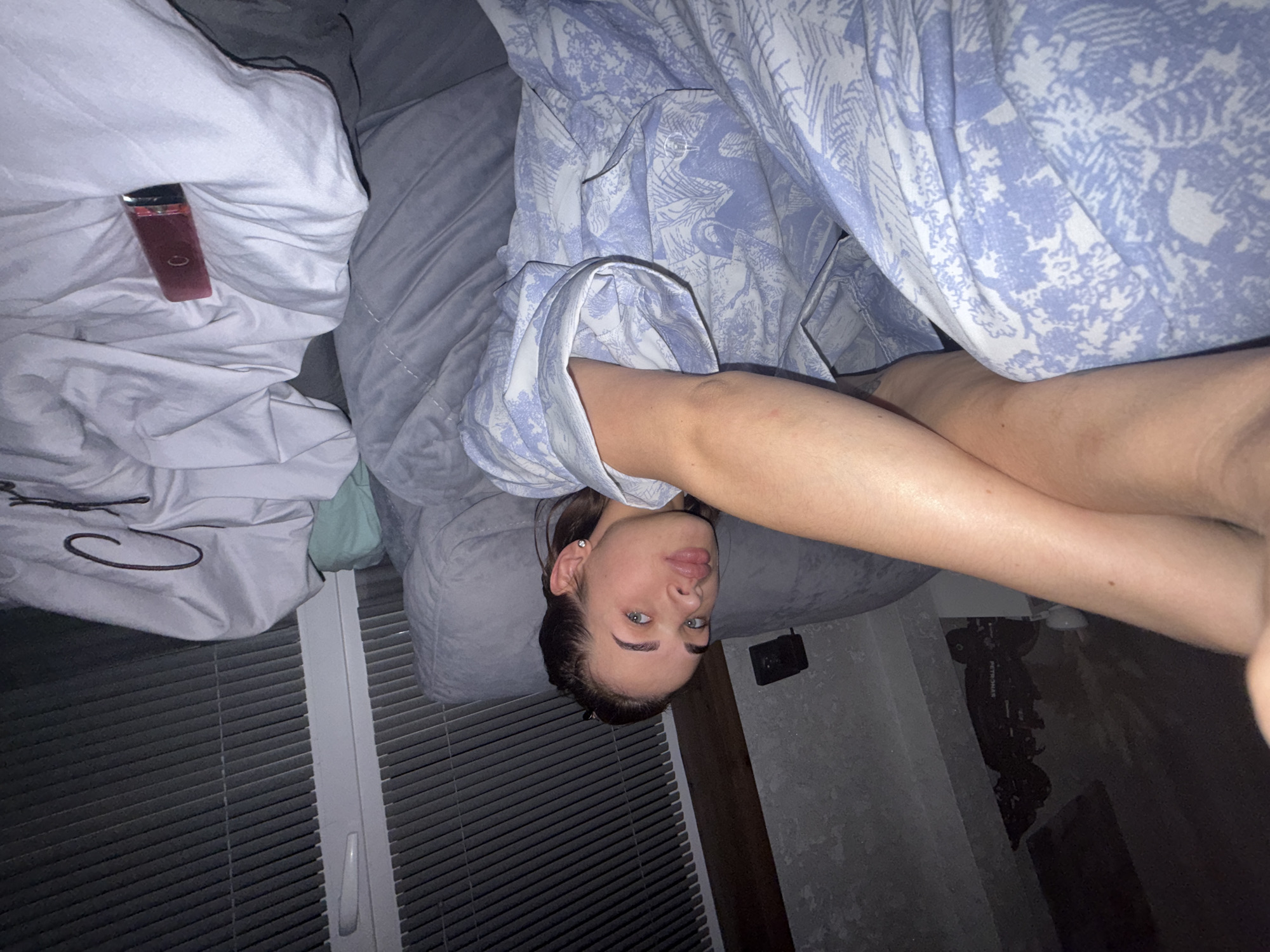 The image shows a woman lying on a bed in what appears to be an indoor setting. She is wearing a light blue and white patterned shirt. The bed is covered with white and gray bedding. A red object is visible on the bedding near the top left corner of the image. Blinds cover a window in the lower left corner. The location is unspecified, and whether it is in the city of Лобаново, Russia cannot be verified from the image. The room's interior suggests a residential setting.