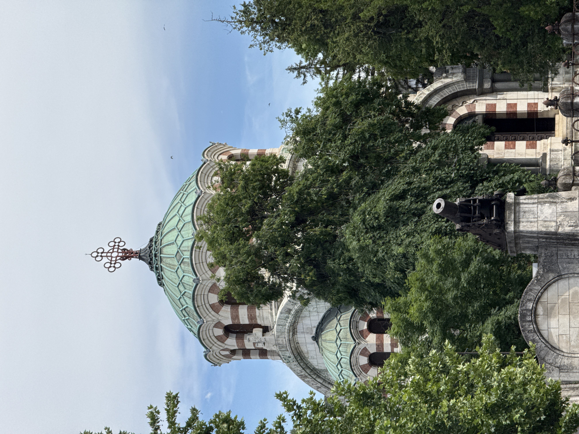 The image shows the exterior of the St. George the Conqueror Pleven Orthodox Church, also known as the Pleven Panorama, located in Pleven, Bulgaria. The church features a prominent green dome topped with a cross. The building's facade is a mix of white stone and red brick detailing around the arched windows and doorways. Large, mature trees with lush green foliage partially obscure the view of the church, creating a contrast between the architecture and the natural environment. In the foreground, to the left of the church entrance, a historical cannon is visible, mounted on a stone pedestal. The sky is partly cloudy with a few birds flying in the distance. The overall scene depicts a historical and religious landmark surrounded by nature.