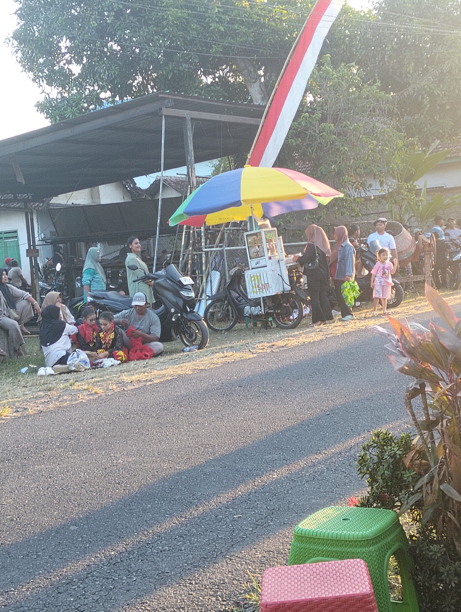 The image depicts a street scene in Sumbersewu, Indonesia, bathed in the warm light of late afternoon. A group of people are gathered on the side of a paved road, some sitting on the grass and others standing. In the background, a building with a corrugated metal roof is visible, alongside several motorcycles and a food cart. A multi-colored umbrella shades the food cart, which appears to be selling snacks or drinks. A tall, striped flag, red and white, is prominently displayed in the upper right portion of the frame, leaning against a tree. The overall atmosphere suggests a casual outdoor gathering, possibly a local event or a marketplace.
