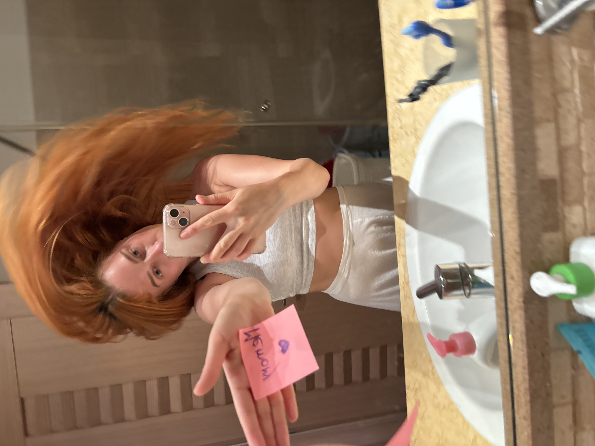 In a bathroom mirror in Budva, Montenegro, a woman with long, red hair, which appears to be in mid-motion or tossed, is taking a self-portrait. She holds a light-colored smartphone with a clear case and a dual-lens camera system in her right hand, pointed towards her reflection. Her face is visible, and she looks towards the phone. She is wearing a light gray ribbed crop top and light-colored lower garments. Her left arm is extended towards the camera, holding a pink sticky note with purple writing, which reads "moM" (backward) and includes a small heart drawn underneath.
The bathroom setting includes a light-colored counter with a white porcelain sink. On the counter, a pink-capped soap dispenser and a chrome faucet are visible. To the right of the sink, a white cylindrical container holds a black toothbrush and a blue toothbrush. Partially visible in the bottom right corner is a white bottle with a green pump. The wall to the left of the woman's reflection features a light-colored, possibly wooden, slatted paneling. To her right, the reflection shows a glass shower enclosure. A white bucket is partially visible behind her right arm in the reflection.