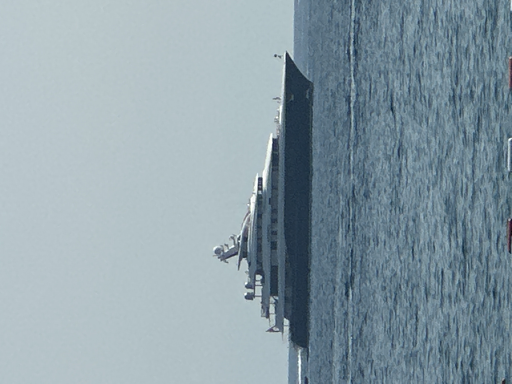 A large, dark-hulled superyacht is sailing on a calm, blue sea under a clear sky. The yacht has multiple decks with white railings and several windows. A prominent superstructure with antennas and a mast rises from the rear of the yacht. The yacht is moving from left to right, leaving a small wake behind it. In the foreground, the water is rippled. The scene appears to be the Bay of Kotor, near Boreti, Montenegro.