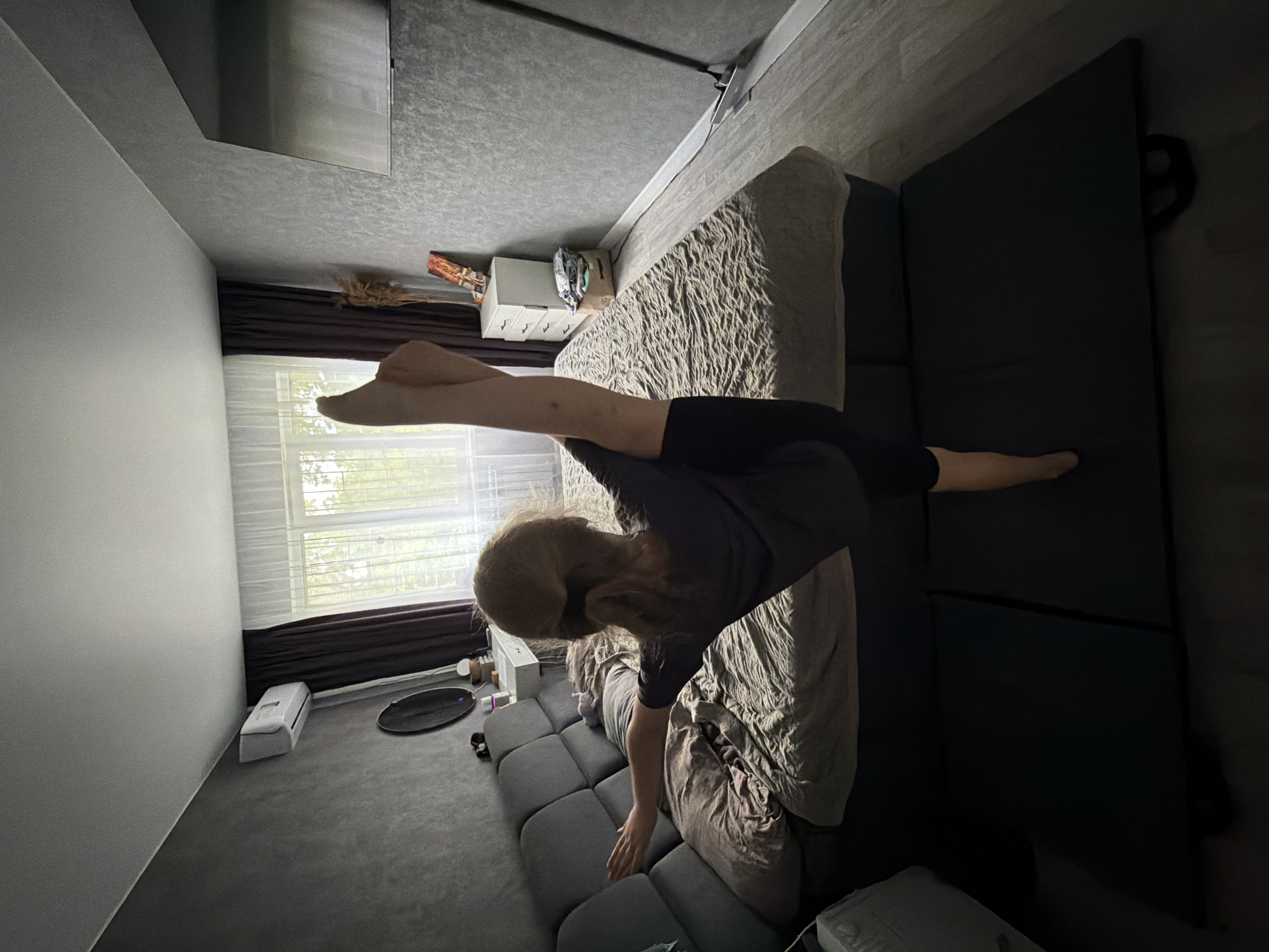 In a bedroom, a person with blonde hair pulled back in a ponytail is performing a vertical split. They are wearing a dark t-shirt and black leggings. Their right leg is extended upwards in front of them, while their left leg is bent at the knee, supporting their stance on a dark exercise mat. The person's left arm is extended towards a gray sectional sofa, with their hand resting on it. The sofa is covered with pillows and a textured bedspread. Behind the sofa, a white dresser with three drawers is visible. On top of the dresser are a framed picture, a vase with dried reeds, and a basket of folded clothes. To the left of the dresser, a large window is covered by sheer white curtains and darker brown drapes. An air conditioning unit is mounted on the wall above the window. To the right of the window, a large flat-screen television is mounted on the textured gray wall. The floor is light gray wood laminate. The lighting in the room is dim, with natural light filtering in through the window and artificial light from the air conditioning unit. The overall impression is that of a residential bedroom being used for athletic training.
