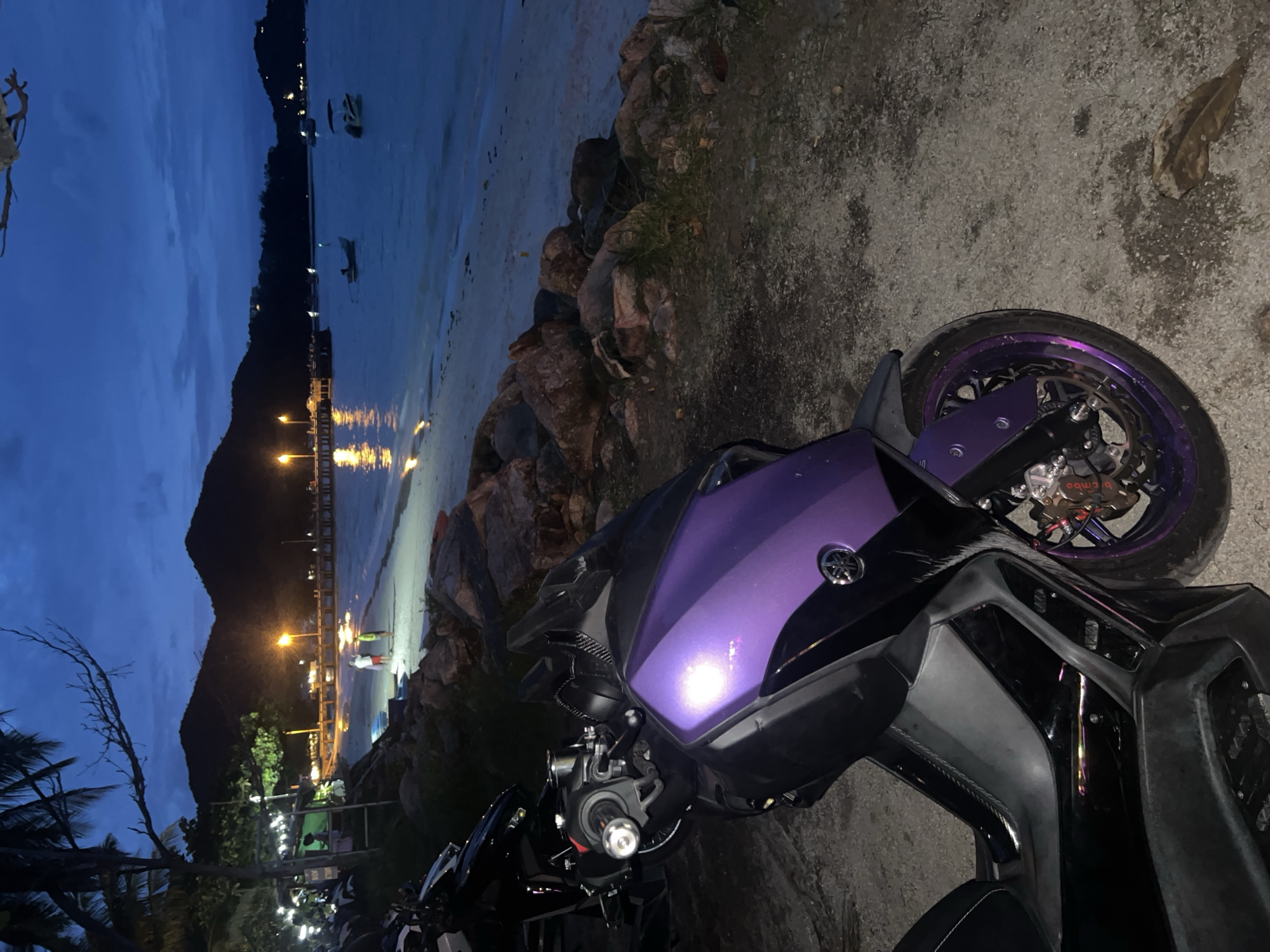 A purple scooter is parked on a dirt path by a rocky shoreline on the island of Ko Pha-ngan, Thailand. In the background, a pier juts out into a body of water, with lights reflecting on its surface. Two people are visible on the beach, and several boats are anchored in the water. The sky is dark blue, indicating dusk or night, with rolling hills in the distance.