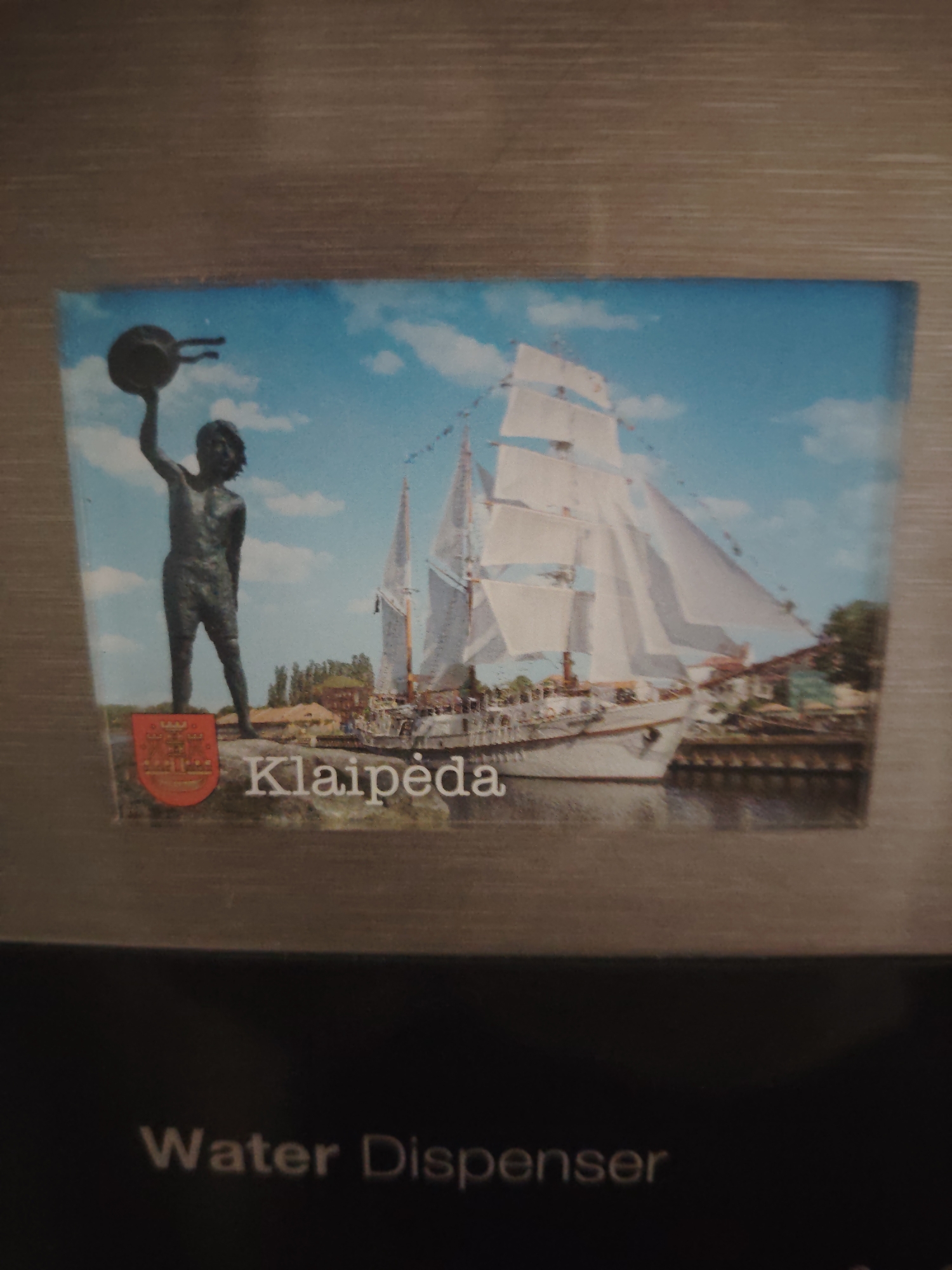 The image is a postcard featuring a scene of a statue of a boy holding a pot above his head, a tall ship with white sails, a body of water, and buildings in the background. The word "Klaipeda" is written in large white letters across the bottom of the image. Below the postcard, the words "Water Dispenser" are visible. The image is taken from a slightly angled perspective and appears to be a photograph of a postcard that is attached to a surface, possibly a refrigerator. The postcard depicts a historical or cultural scene related to the city of Klaipeda. The location of this postcard is not within Huntingdonshire, United Kingdom, but rather related to the city of Klaipeda, Lithuania.