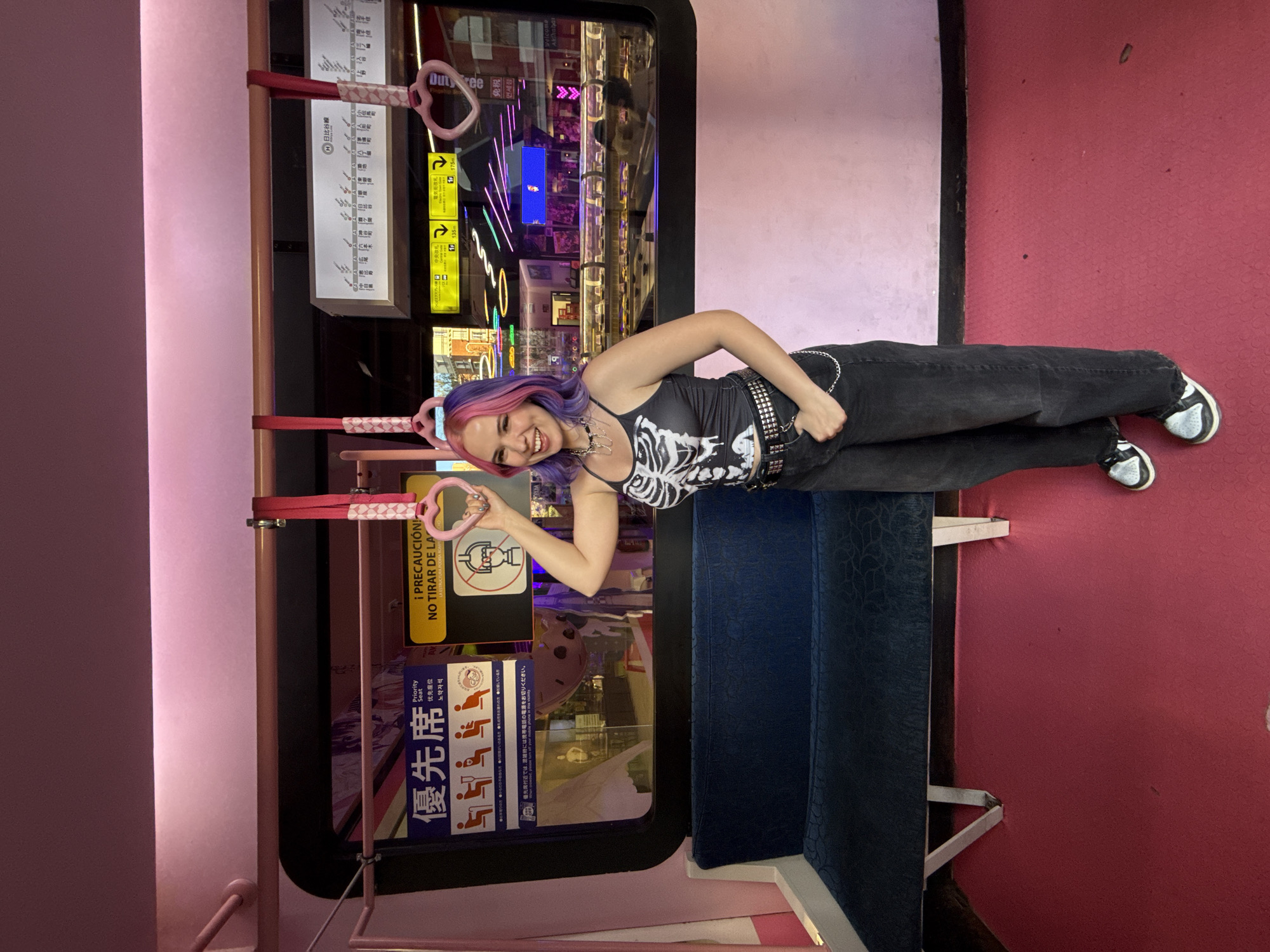 A full shot captures a young woman on what appears to be public transportation, possibly a train. She is fair-skinned with hair dyed purple and pink, adorned with a matching headband. She wears a black tank top with a skeleton print, dark jeans, and sneakers. Her accessories include a studded belt and a necklace. She is smiling and posing with a handrail in the shape of a heart.
The interior of the transportation vehicle is decorated in shades of pink and red, with pink walls and a red floor. There is a blue padded bench seat nearby. The window behind the woman displays advertisements and signs written in Japanese, indicating a location likely in Japan, not Madrid, Spain. Among the signs are warnings in Spanish ("¡PRECAUCIÓN! NO TIRAR DE LA") and markings for "Priority Seat." The vehicle also features a map of the Hibiya Line subway route.