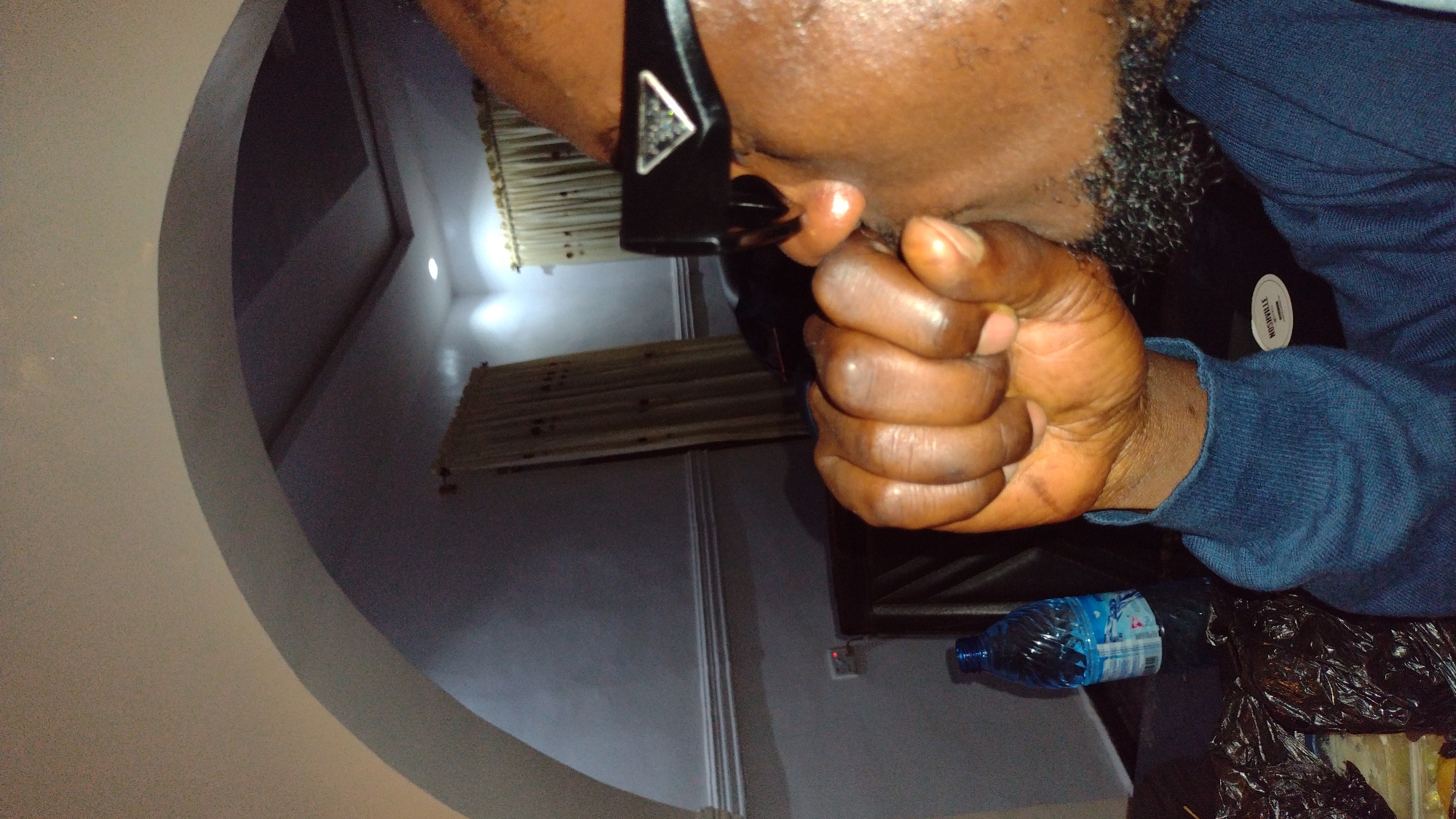 The image shows a close-up of a person's face and hand in what appears to be an indoor setting. The person is wearing dark-rimmed glasses with a triangular logo on the side. Their hand is clenched into a fist, held near their face, partially obscuring their cheek and beard. The person is wearing a dark blue long-sleeved shirt.
In the background, there is an arched architectural feature, possibly a doorway or alcove. Further back, curtains are visible, along with a ceiling with recessed lighting. On a surface in the foreground, to the left, is a clear plastic bottle of water with a blue label. There are also dark plastic bags, suggesting trash, on the surface. The lighting in the room is dim, with light sources seemingly coming from above and from an illuminated outlet on the wall.
There is no information in the image that allows for the determination of the location being in Jos, Nigeria.