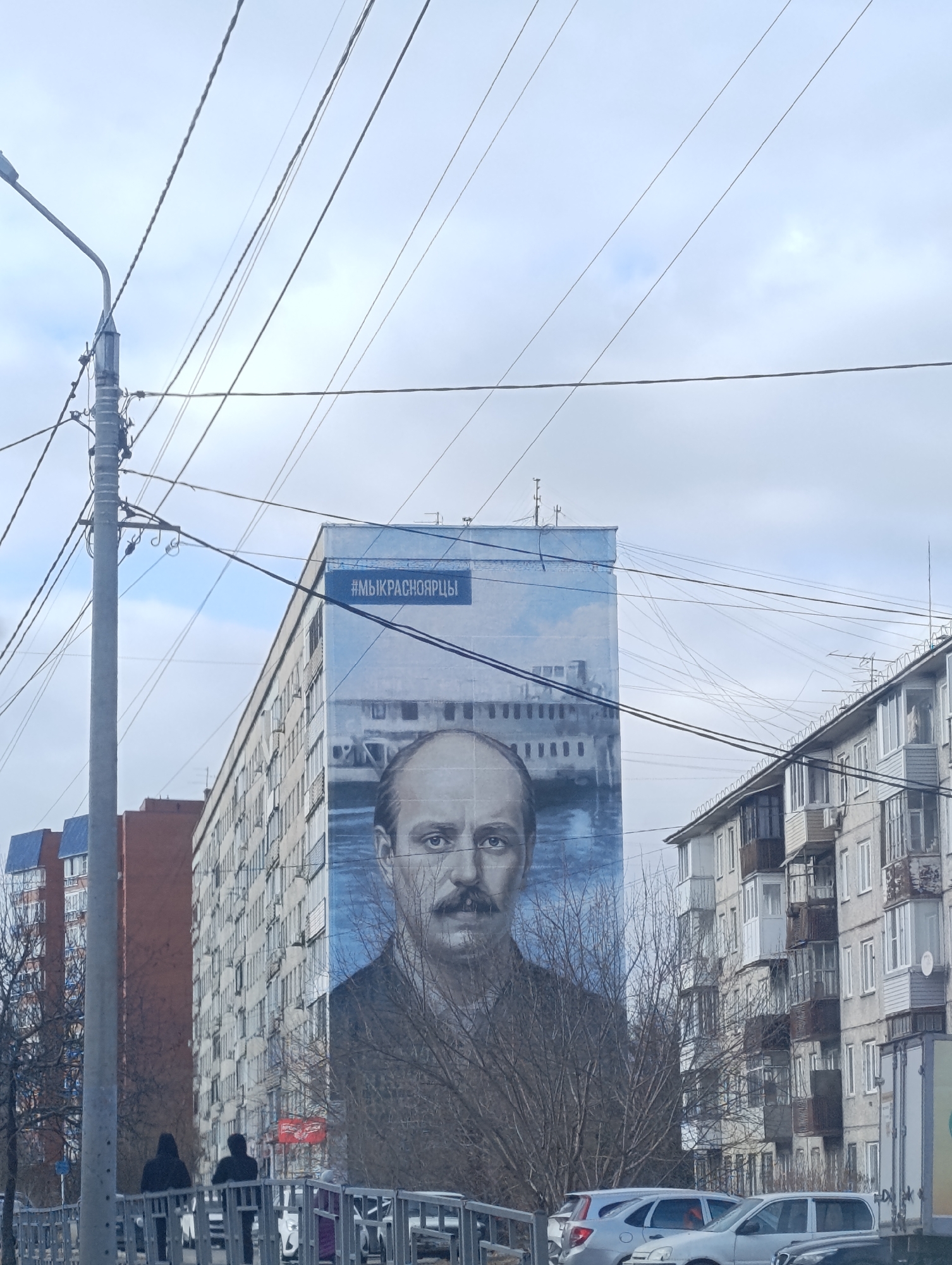 An urban street scene in Krasnoyarsk, Russia, unfolds under an overcast, cloudy sky. The dominant feature is a monumental mural painted on the entire side of a tall, multi-story residential apartment building on the left. The mural vividly portrays a bald man with a prominent mustache, his gaze directed forward with a serious or contemplative expression. Behind his head, a large steamboat is depicted navigating a body of water, beneath a sky with soft, diffused clouds. A blue banner at the very top of the mural proudly displays the white Russian text `#МЫКРАСНОЯРЦЫ`, which translates to `#WEAREKRASNOYARSKRESIDENTS`.
In the foreground, a tall utility pole with a street lamp stands prominently on the left, from which numerous power lines extend across the upper portion of the frame. A metal railing separates the viewer from a street where several cars are visible, including silver sedans and a hatchback, indicating a daytime setting. Two silhouetted figures are seen walking near the railing on the lower left, and another person is partially visible among the parked vehicles on the right. To the right of the mural building, another, older apartment block with numerous balconies is visible, with bare trees suggesting a colder season like late autumn or early spring. Additional residential buildings, including a distinct red-brown structure, are discernible in the background on the left.