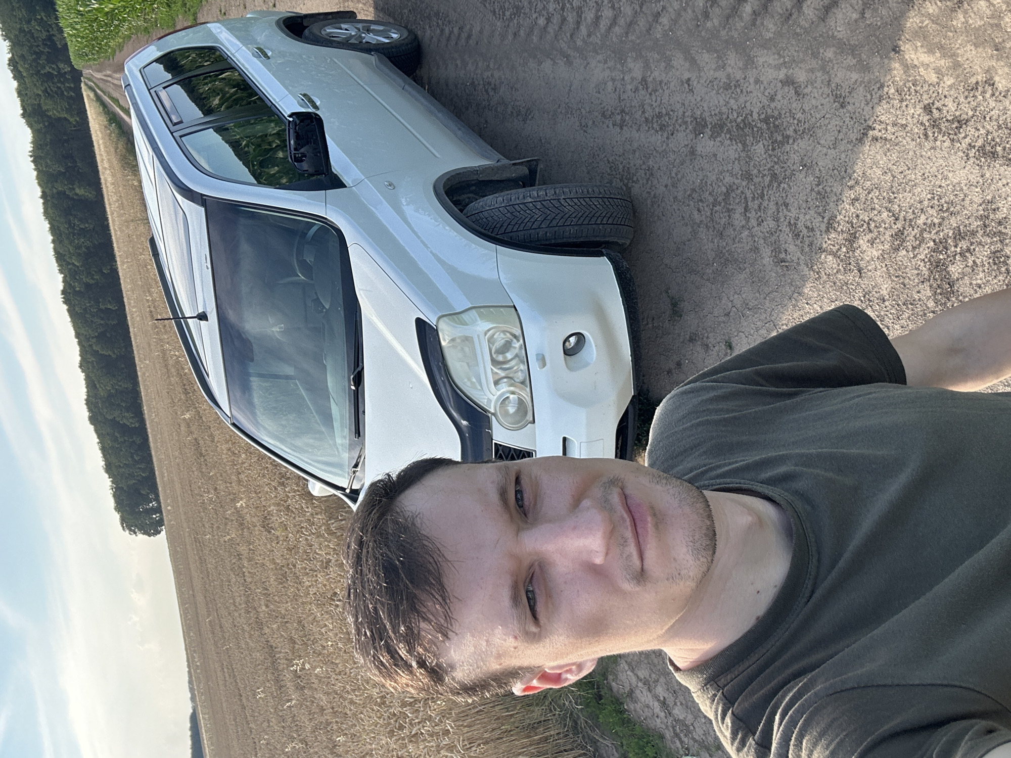 A man is taking a selfie in front of a white SUV parked on a dirt road in Makiv, Ukraine. The man is wearing a dark green t-shirt and has short brown hair. The SUV is a Nissan X-Trail, and it is parked on a dirt road that appears to be in a rural area. To the left of the road is a field of dry wheat, and to the right is a field of green corn. In the background, there is a line of trees and a blue sky with some clouds. The sun is shining, and the lighting suggests it is daytime.