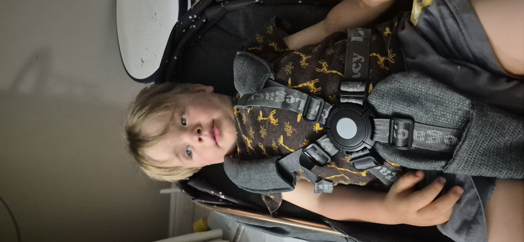 A young Caucasian child with blonde hair and blue eyes sits in a stroller, looking to the left. The child is wearing a brown t-shirt with a pattern of yellow giraffes and the child is secured by a five-point harness. The stroller has dark fabric with a white star pattern. The background is out of focus and appears to be an indoor setting with a plain wall. The lighting is dim.