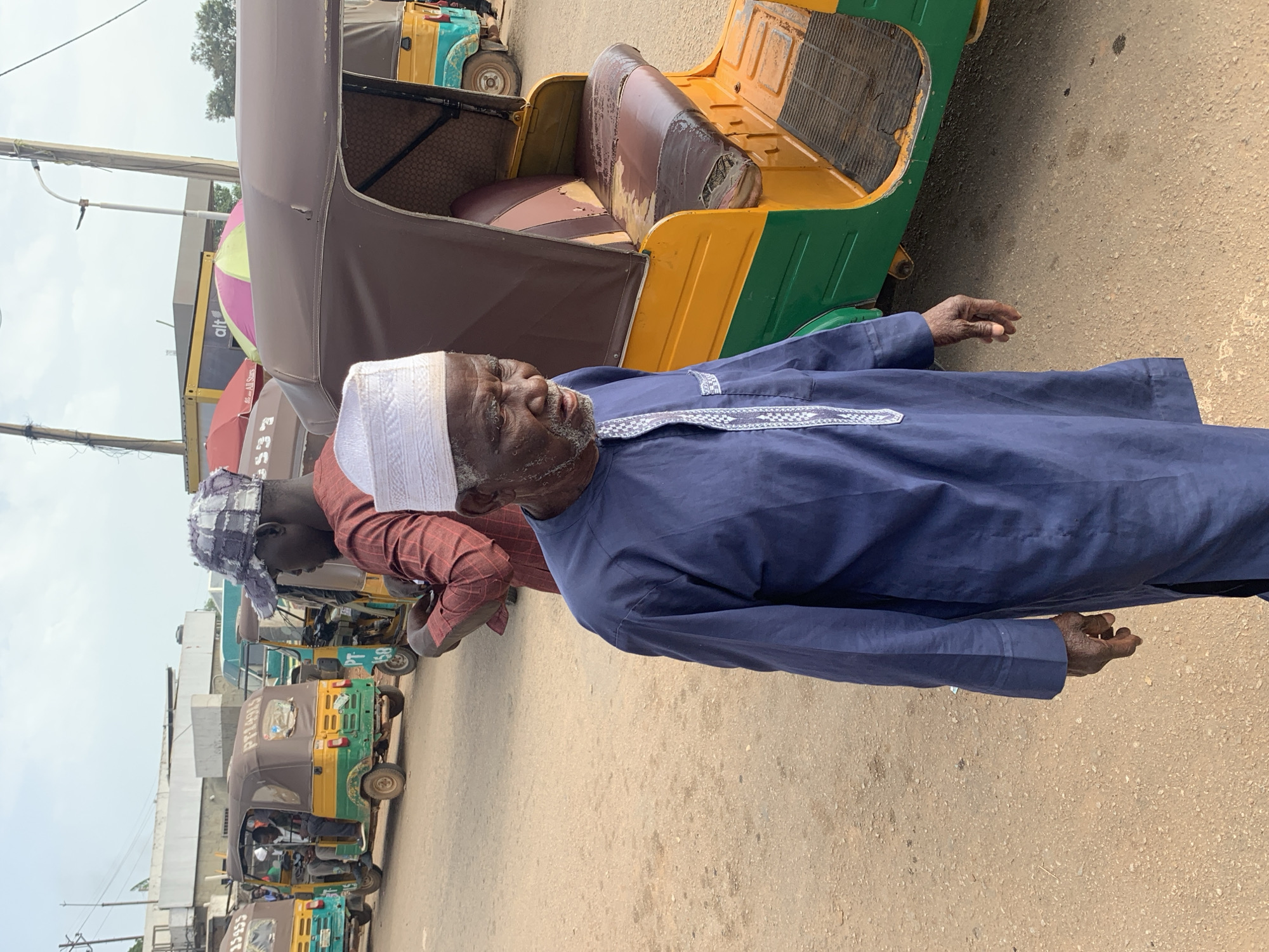 This is a medium shot of an elderly man in a blue robe and white cap standing in a dusty area, likely a street or market, in Gura Top, Nigeria. Behind him, several colorful auto-rickshaws, also known as "keke napep," are parked. The auto-rickshaws are green and yellow with brown canopies. One auto-rickshaw directly to the right of the man has a worn brown leather seat and a visible footboard. The man is looking slightly down and to his right, with his left hand resting at his side. His skin appears weathered, and he has a beard. The background shows more auto-rickshaws and buildings, along with utility poles and wires. The overall scene depicts a common mode of transportation and daily life in this Nigerian city.