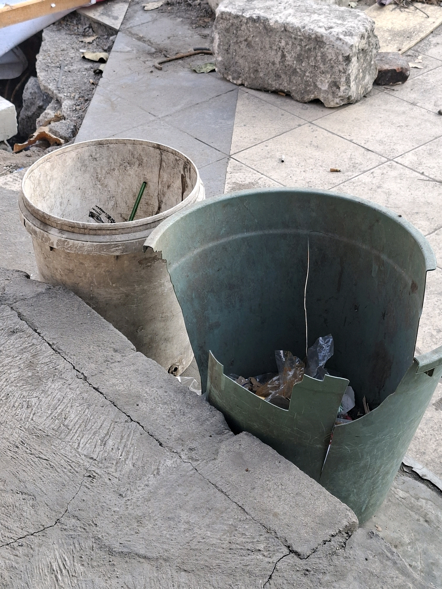 Two broken plastic trash bins are situated on a cracked concrete surface next to some pavement. The bin on the left is a lighter color and appears less damaged. The bin on the right is dark green, significantly cracked, and contains some crumpled plastic and debris. In the background, broken concrete and pavement suggest a construction or demolition site. The environment is outdoors, with natural lighting. The overall scene depicts urban neglect and disrepair, a common sight in certain areas of Surabaya, Indonesia, where infrastructure may be aging or undergoing renovation.
