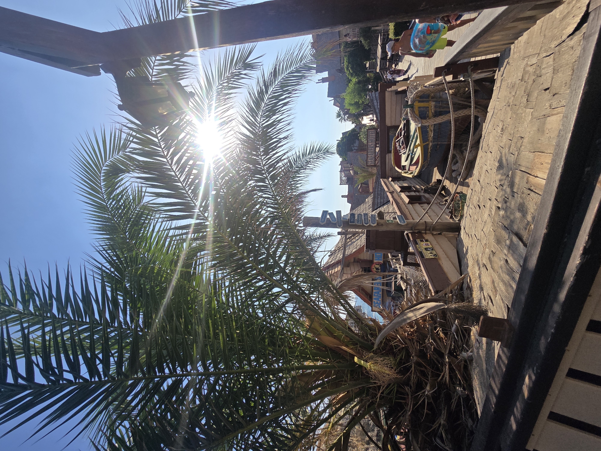 The image depicts a sunny outdoor scene, likely in a resort or themed area, with a prominent palm tree in the foreground casting shadows. Sunlight streams through the palm fronds, creating lens flare. In the background, there is a collection of wooden structures that resemble a village or a pirate-themed attraction, complete with thatched roofs and weathered wood. A small boat is prominently displayed on a wooden deck, adorned with ropes and fishing nets, suggesting a maritime or pirate theme. Several signs with directional arrows and text are visible, indicating different locations. A person wearing a white cap and yellow shorts is walking on a wooden walkway, carrying a blue and red inflatable. The architecture and the presence of the sea in the background suggest a coastal location. The scene evokes a holiday or leisure atmosphere. Based on the architectural style and the information about the location, this scene is likely from Popeye Village in Mellieħa, Malta, which is a film set built for the 1980 film "Popeye" and now operates as a tourist attraction.