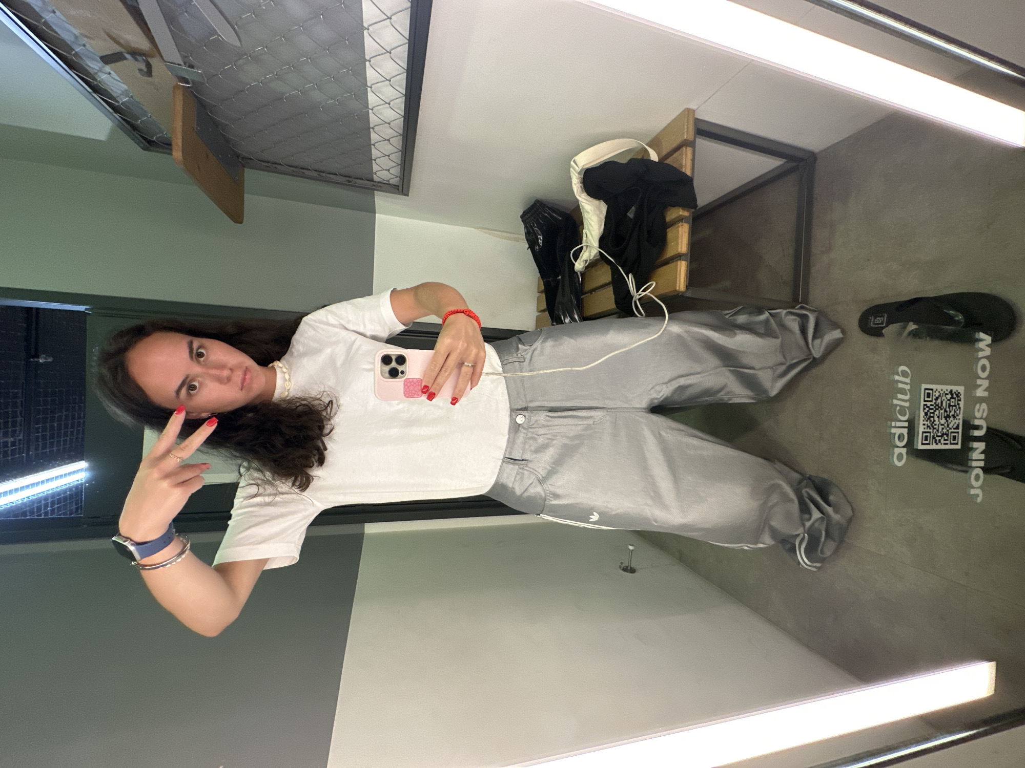 A young woman is posing in what appears to be an Adidas store fitting room. She is wearing a white t-shirt and baggy silver pants with an Adidas logo. She is holding a pink cell phone in her right hand and making a peace sign with her left. Other clothing items are visible on a rack in the background. The fitting room has a wooden bench, and the floor has Adidas branding with a QR code. The store is in Pattaya City, Thailand.