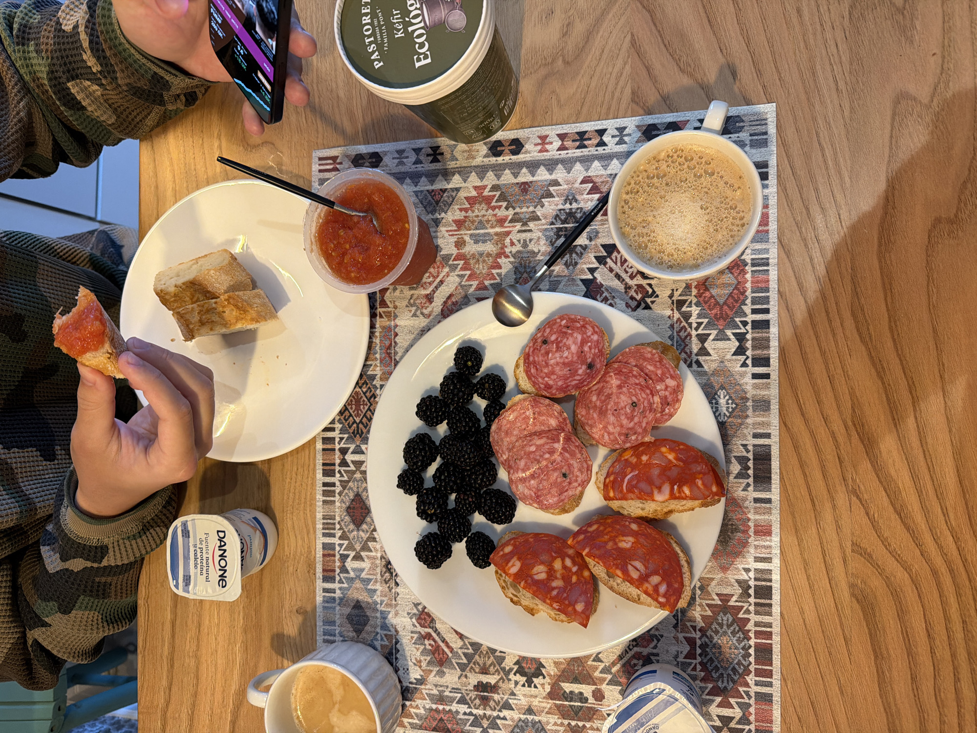 The image shows a table setting with various food items. There are two people present, one holding a phone and the other holding a piece of bread with tomato spread. A plate contains bread, salami, and blackberries. Other items include a container of kefir, a cup of coffee, a small container of tomato spread, and a container of Danone yogurt. The table has a placemat with a patterned design. The table appears to be wooden. The location is not discernible from the image.
