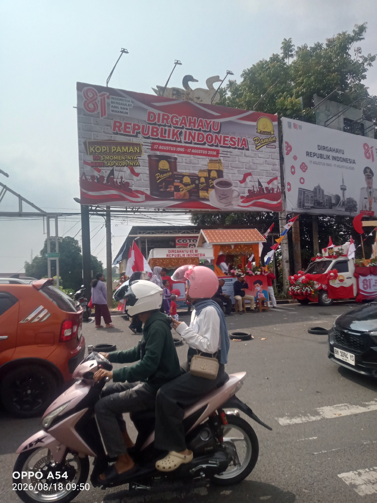 A street scene in Jambi, Indonesia, features a parade or celebration related to Indonesian Independence Day. A large billboard displays "DIRGAHAYU REPUBLIK INDONESIA 17 AGUSTUS 1945 - 17 AGUSTUS 2026" with celebratory graphics. Another billboard prominently advertises "KOPI PAMAN PAS MOMEN-NYA MAN TAP KOPI-NYA."
In the foreground, two individuals on a pink scooter are visible, both wearing helmets. The scooter is moving on an asphalt road with painted white lines. A black car with license plate "BH 1094 YS" is to the right. An orange car is parked on the left side of the road.
In the mid-ground, a small decorated float with red and white decorations and flags is parked, with people gathered around it. A few people are standing and walking on the roadside. Trees and utility poles are also present. The sky is clear and bright. The date stamp "OPPO A54 2026/08/18 09:50" is visible in the lower left corner.