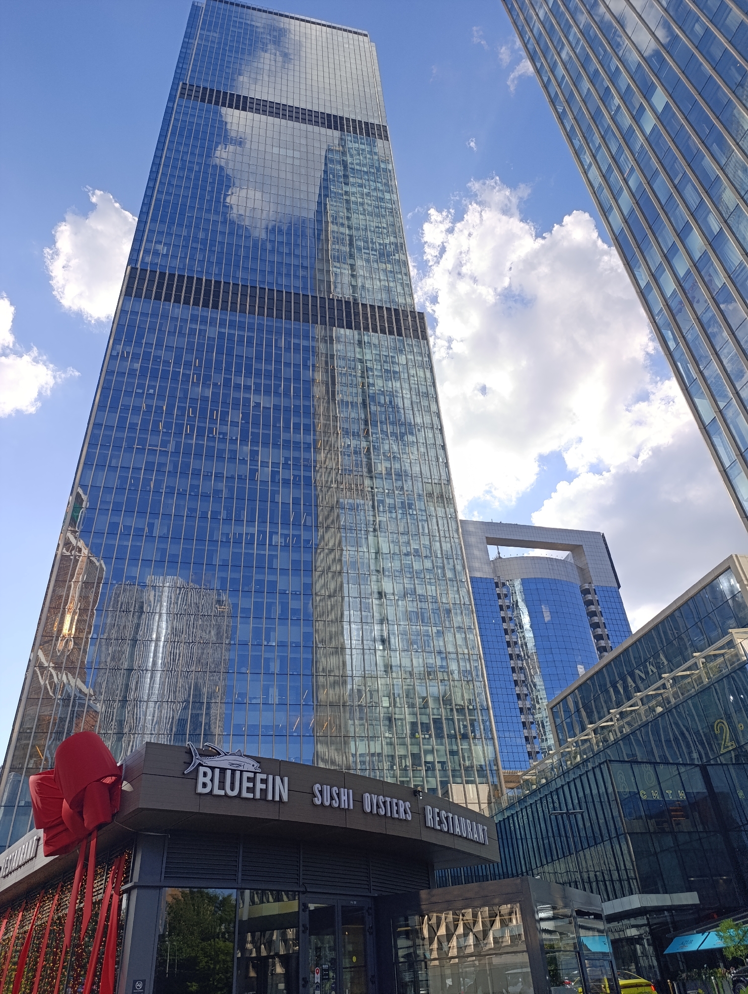 The image captures a vibrant daytime urban landscape in Moscow, Russia, dominated by towering modern glass skyscrapers. The perspective is angled upwards, highlighting the impressive height and reflective surfaces of the buildings against a bright blue sky dotted with fluffy white clouds.
The central and most prominent skyscraper features a facade of blue-tinted glass panels, reflecting the sky, clouds, and distorted images of adjacent structures. Its design incorporates regular horizontal dark bands. To the right, another glass building with similar reflective qualities angles into the frame, showcasing its own grid-like window pattern. In the background, a distinctive skyscraper with a large, arched opening near its summit is visible, adding to the architectural diversity.
In the foreground, at the base of one of the main buildings, is a commercial establishment. A prominent sign displays "BLUEFIN" in white letters with an accompanying fish logo. Below this, the words "SUSHI OYSTERS RESTAURANT" indicate its culinary offering. The restaurant's facade is decorated with a large, festive red bow and hanging ribbons, suggesting a special event or holiday theme.
Visible text on other buildings includes "VANKA," "2° СИТИ" (2° CITY), and "СИТИ" (CITY), reinforcing the context of a modern urban development like Moscow City. The scene is bright and sunny, contributing to the clear reflections and vibrant colors. No people are distinctly visible, leaving the focus on the architectural grandeur and the interplay of light and reflection on the glass surfaces.