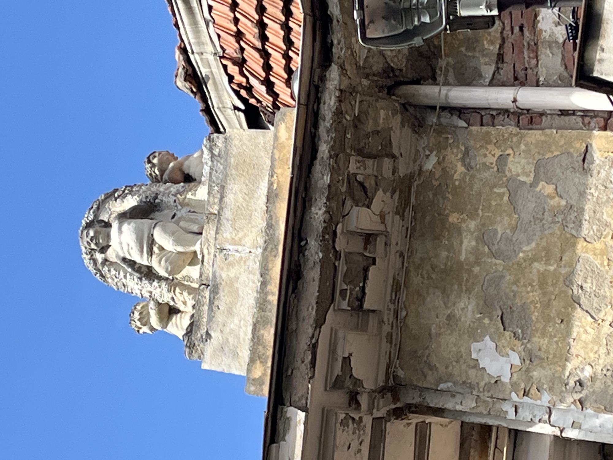 The image displays a detailed view of the upper portion of an old building in Sofia, Bulgaria, under a clear blue sky. On the left side, against the bright sky, rests a prominent, light-colored stone sculpture or statue. This main figure appears to be a classical-style woman, possibly seated or reclining, with another smaller figure, potentially a child or cherub, on or near her. Below this, a second, smaller bust-like sculpture is partially visible on the same weathered stone ledge.
To the right of the sculptures, the building's facade shows significant signs of age and disrepair. Large sections of light-colored paint have peeled away, revealing rough plaster and red brickwork beneath. A terracotta-tiled roof is visible along the top right edge, with its distinct pattern of overlapping tiles. A white pipe or conduit runs horizontally along the weathered wall, and a dark, possibly metallic, fixture is attached to the upper part of the building, near the roofline. The bright, even lighting and crisp shadows indicate it is daytime with sunny weather. No visible text is present.