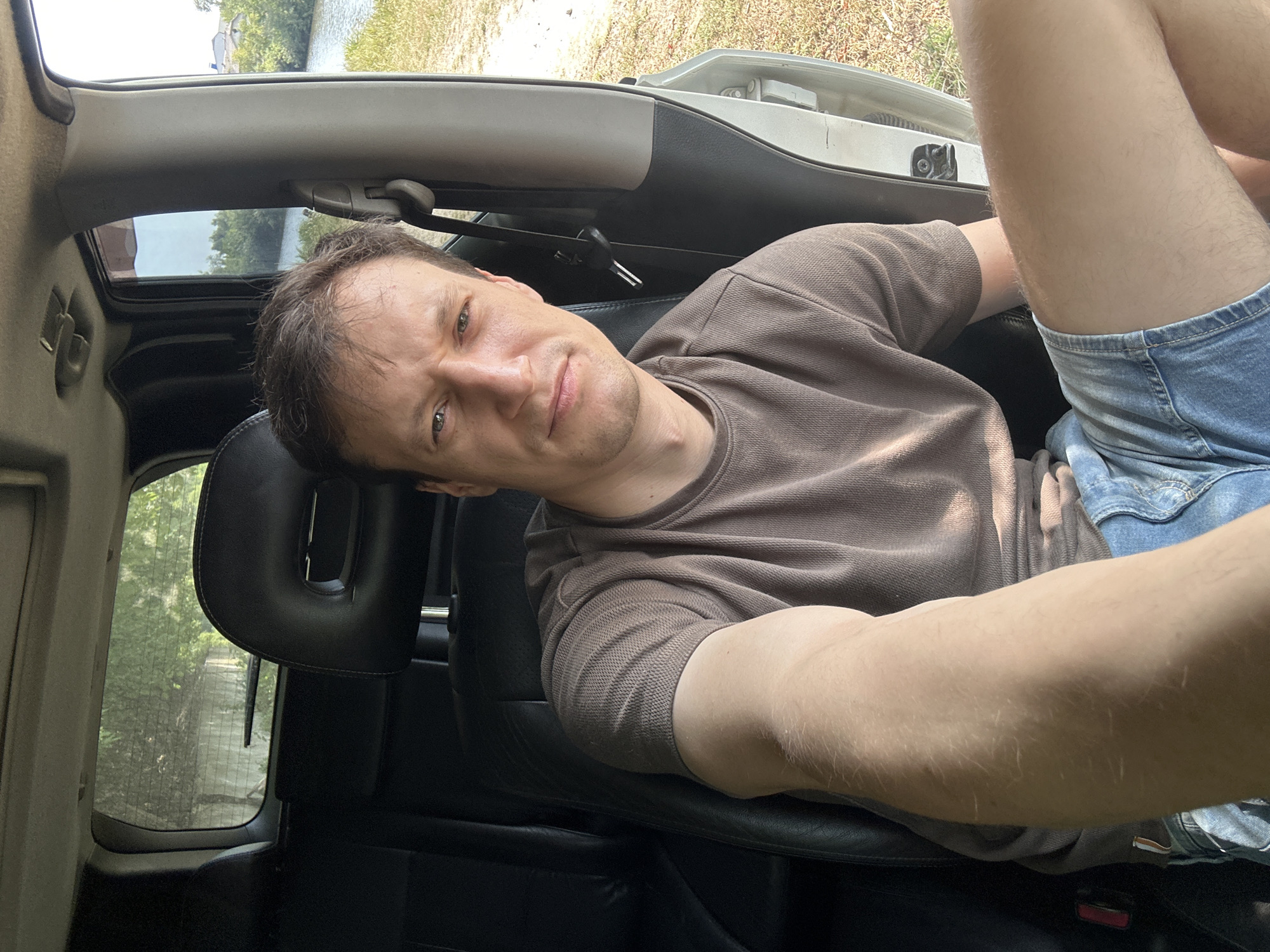 A man with short brown hair, wearing a brown t-shirt and denim shorts, is sitting in the driver's seat of a car. He is looking directly at the camera. The car's interior is visible, including black leather seats and a sunroof. Outside the car, there is a body of water to the right, with grassy banks and trees in the background. The scene appears to be in a rural or semi-rural area, possibly near Makiv, Ukraine, given the context. The lighting suggests it is daytime.