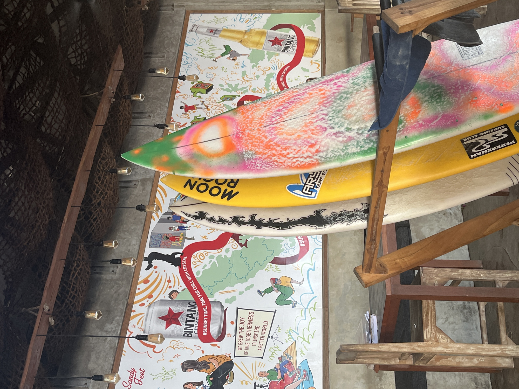The image captures a vibrant scene in North Kuta, Indonesia, likely at a surf shop or beachside cafe. In the foreground, three surfboards stand vertically, leaning against a wooden rack. The closest surfboard is predominantly white with a chaotic, colorful spray paint design in pink, orange, and green. To its left, a bright yellow surfboard displays text that appears to be "MOON" in large black letters. The third, partially visible surfboard on the far left, is white with black decorative markings.
Behind the surfboards, a large, colorful mural adorns the wall. The mural features stylized illustrations, including a prominent Bintang beer can and bottle, a world map outline, and various figures engaged in activities like dancing and surfing. Text within the mural includes "Sandy Feet," "#SUNSET TIME, TIME FOR CHILL WITH CRYSTAL," and "WE BREW THE JOY OF TRUE TOGETHERNESS TO INSPIRE A BETTER WORLD."
The environment suggests an indoor space with a rustic, open-air feel. Exposed wooden beams crisscross the ceiling, from which several bare Edison-style light bulbs hang, illuminating the scene. The wall behind the mural is a textured concrete or stucco material. In the lower left corner, a dark wooden table with chairs is visible, suggesting a seating area. Papers are stacked on the table, and one chair is pushed in, indicating recent use. The overall impression is a casual, beach-themed establishment.