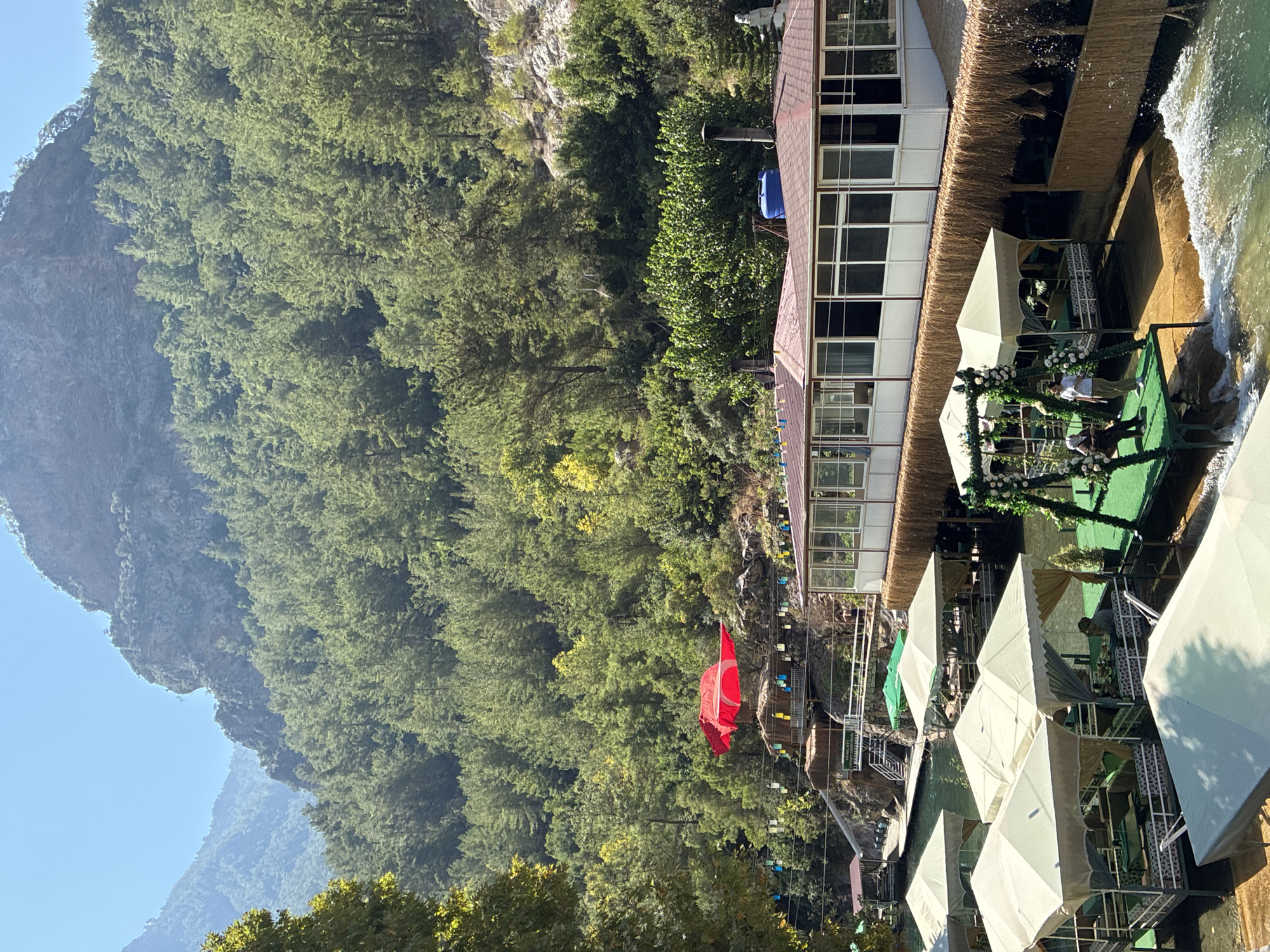 In Alanya, Turkey, this image captures a scenic riverside dining establishment nestled within a verdant, mountainous landscape. The foreground features several open-air dining platforms partially covered by white awnings, situated on the water's edge. These platforms are equipped with tables and seating, with a few patrons visible. A decorative swing adorned with flowers is present. Behind the platforms, a long, white building with a red roof and numerous windows extends horizontally. Further back, a large red flag with a white crescent and star, the Turkish flag, is prominently displayed, billowing in the wind, positioned above a thatched-roof structure. The background is dominated by a steep, tree-covered mountain slope, indicating a natural, possibly remote, location. The water is clear and appears to be flowing. The overall atmosphere suggests a tranquil outdoor recreational and dining area.