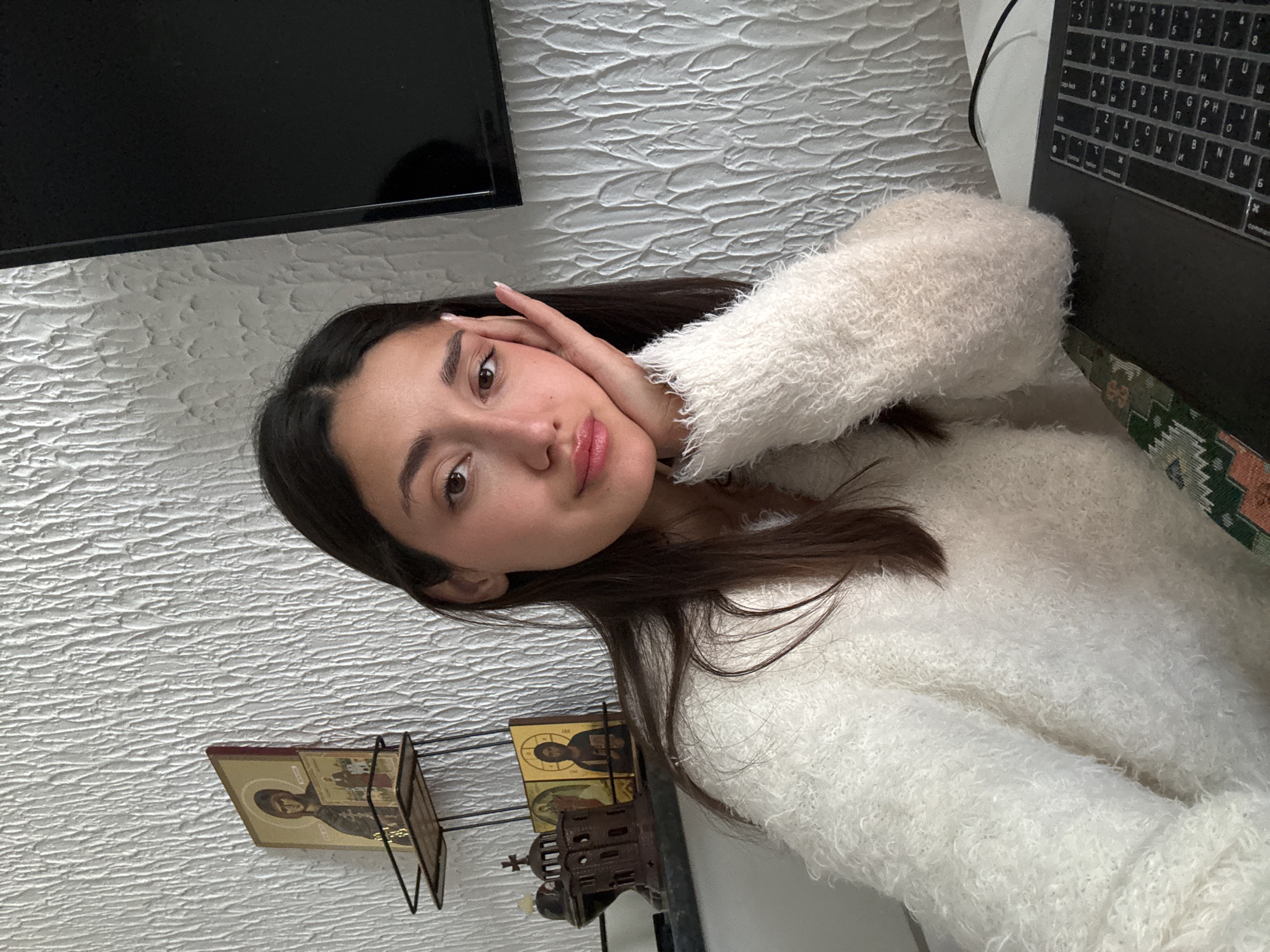 An indoor scene in Sutomore, Montenegro, depicts a young adult female seated at a white desk in front of a textured, light-colored wall. The female, centered in the frame, has dark hair parted centrally and wears a cream-colored, fuzzy knit sweater. Her left hand, with manicured nails, rests against her left cheek. She looks directly at the camera with a neutral expression. To her left, against the wall, a small dark-framed multi-tiered shelf displays religious icons: a larger rectangular icon on the upper shelf, and two smaller icons and a brown miniature church-like structure with a cross on the lower shelf. To her right, a large dark rectangular screen, possibly a television or monitor, is visible. In the lower right foreground, a dark laptop computer with a keyboard showing both Latin and Cyrillillic characters is open on the desk, with a dark cable extending from it. A patterned fabric cushion or upholstery in green, orange, and cream geometric designs is visible beneath the female's left arm in the lower-left corner.