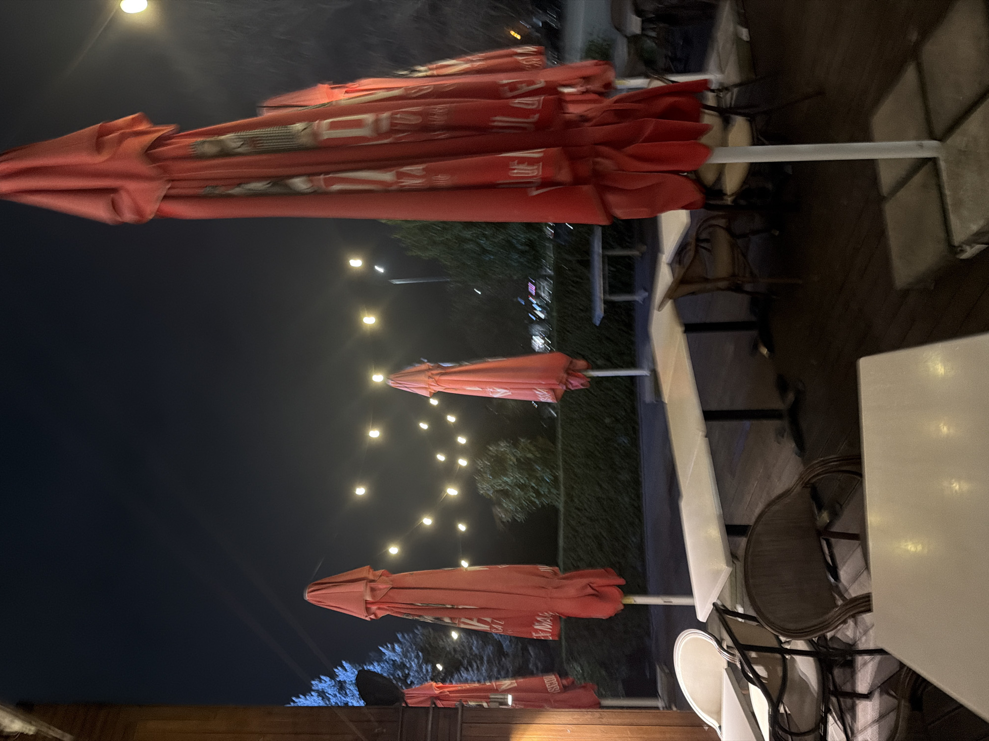 The image depicts an outdoor seating area at night, likely a restaurant or cafe in Plovdiv, Bulgaria. Several red, closed umbrellas are prominent in the foreground and middle ground, possibly bearing brand logos. In the background, strings of lights illuminate trees and foliage. Seating arrangements, including tables and chairs, are visible in the foreground and along the right side of the image. The scene suggests a quiet evening atmosphere in a commercial outdoor space.