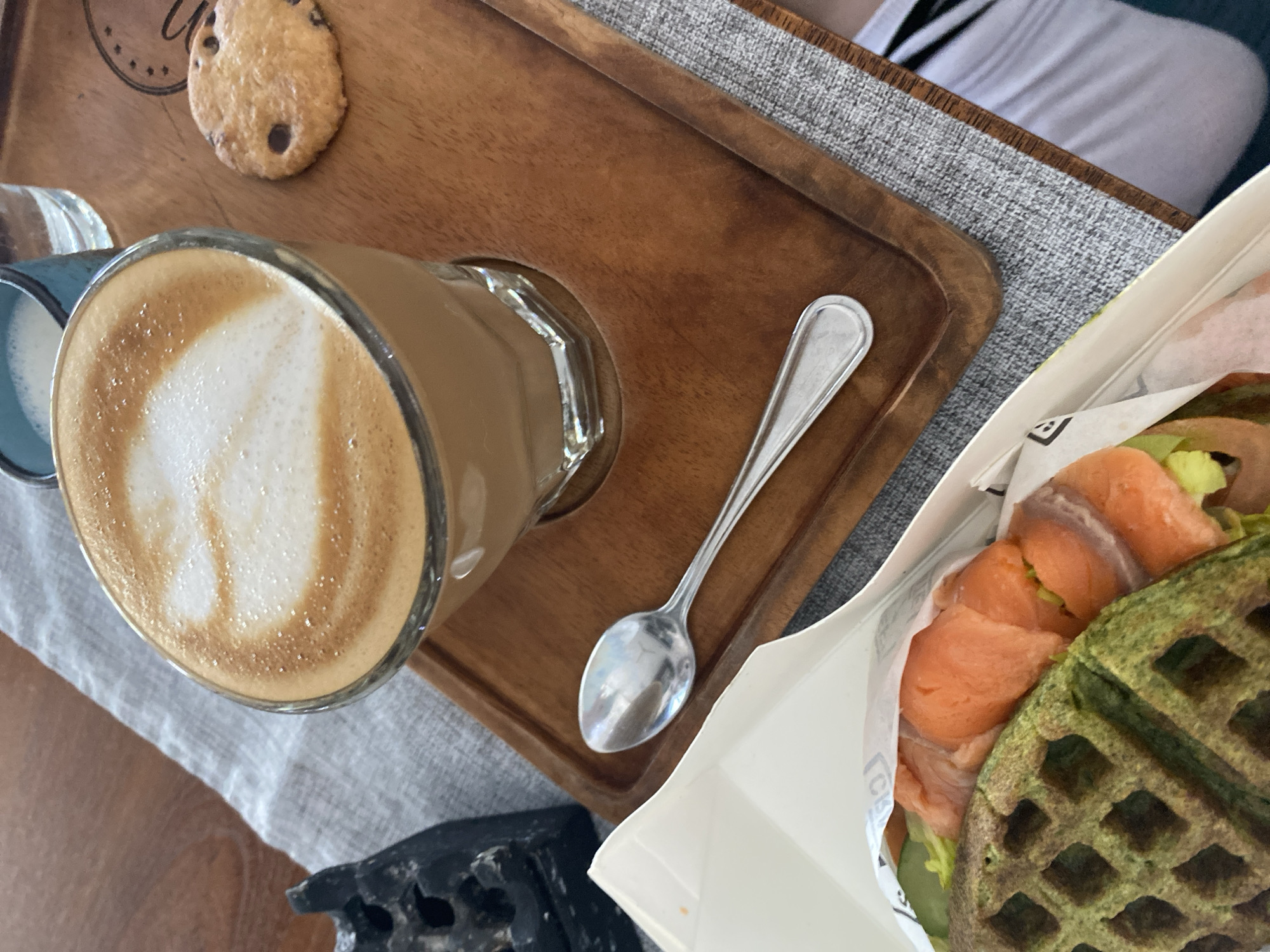 A top-down view shows a wooden tray holding a latte in a glass mug, a small cookie, and a silver spoon. Next to the tray, a white container holds a green waffle sandwich with salmon and lettuce. A person's leg wearing white socks with black stripes is partially visible in the background. A grey table linen is visible under the tray. A small decorative black object is visible at the bottom left corner. The scene is set in Kuta Selatan, Indonesia.