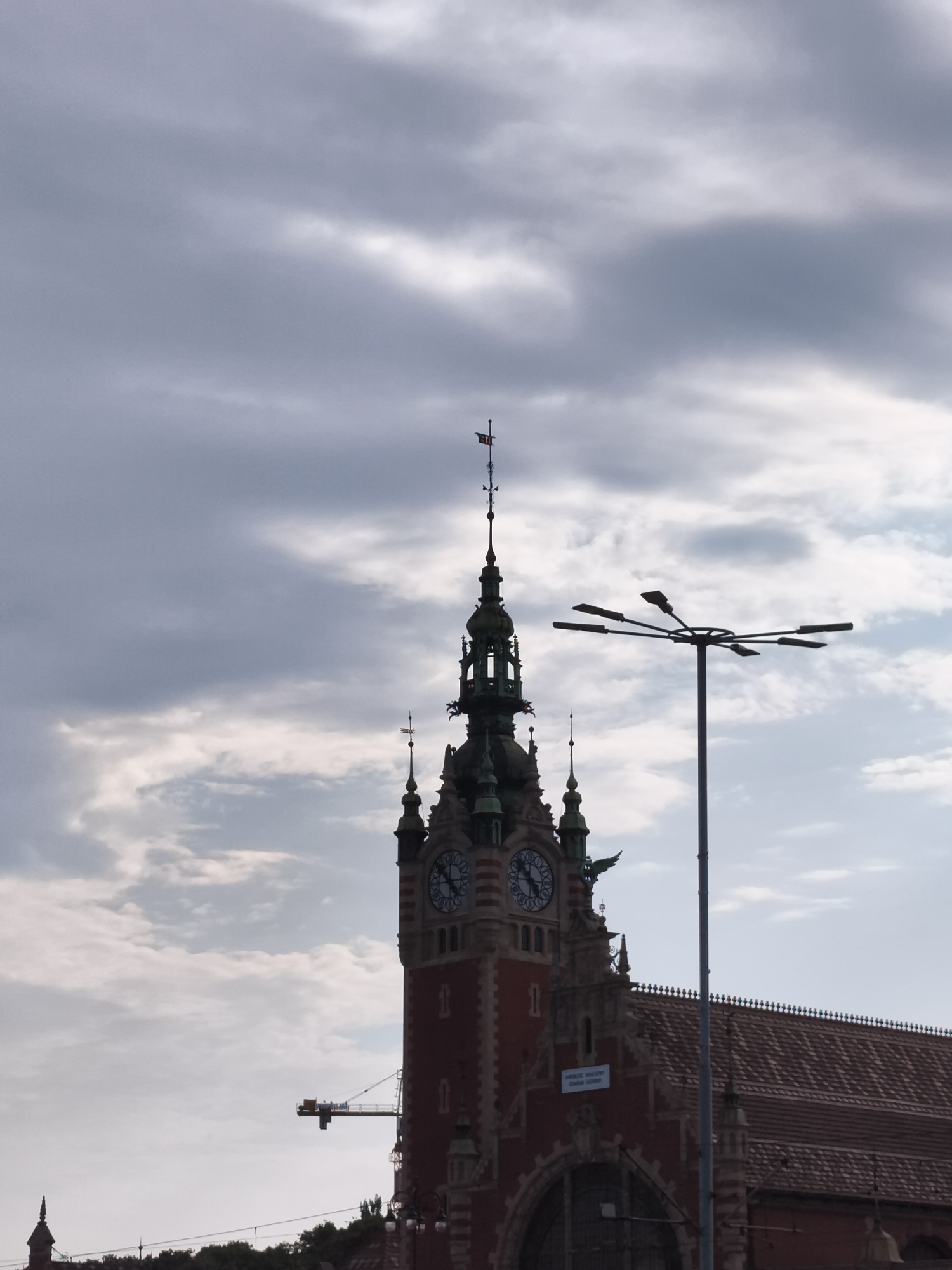The image shows the historic Gdańsk Główny railway station in Gdańsk, Poland, under a cloudy sky. The station's prominent red brick clock tower, with its Gothic Revival architecture and ornate details, is a central focus. The tower features two large clock faces. To the right of the tower, a modern street lamp with multiple arms stands silhouetted against the sky. In the background, a construction crane is visible near the station buildings, suggesting ongoing work. The roof of the station, covered in reddish-brown tiles, is also visible. The scene appears to be captured during daylight, but the overcast conditions create a somewhat somber atmosphere. There are no people or animals clearly discernible in the image.